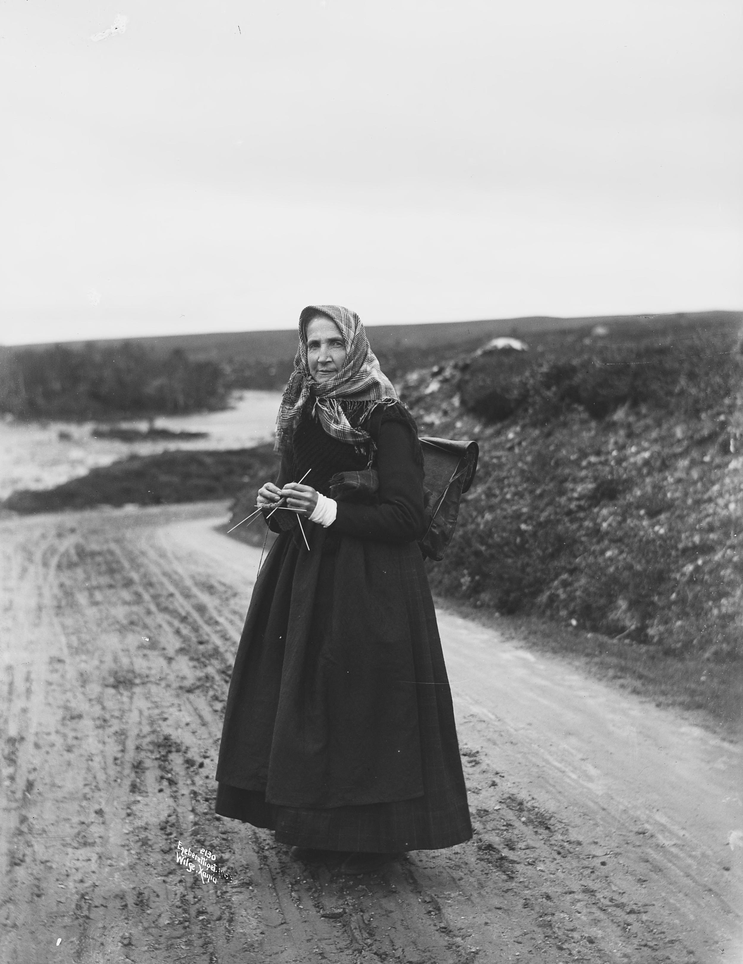 Svart- kvitt foto. Svartkledd kvinne med skreppe står midt i bilde med strikketøy