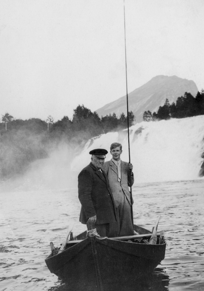 To menn med fiskeutstyr i ein robåt i ei elv framfor ein foss, med fjell i bakgrunnen.