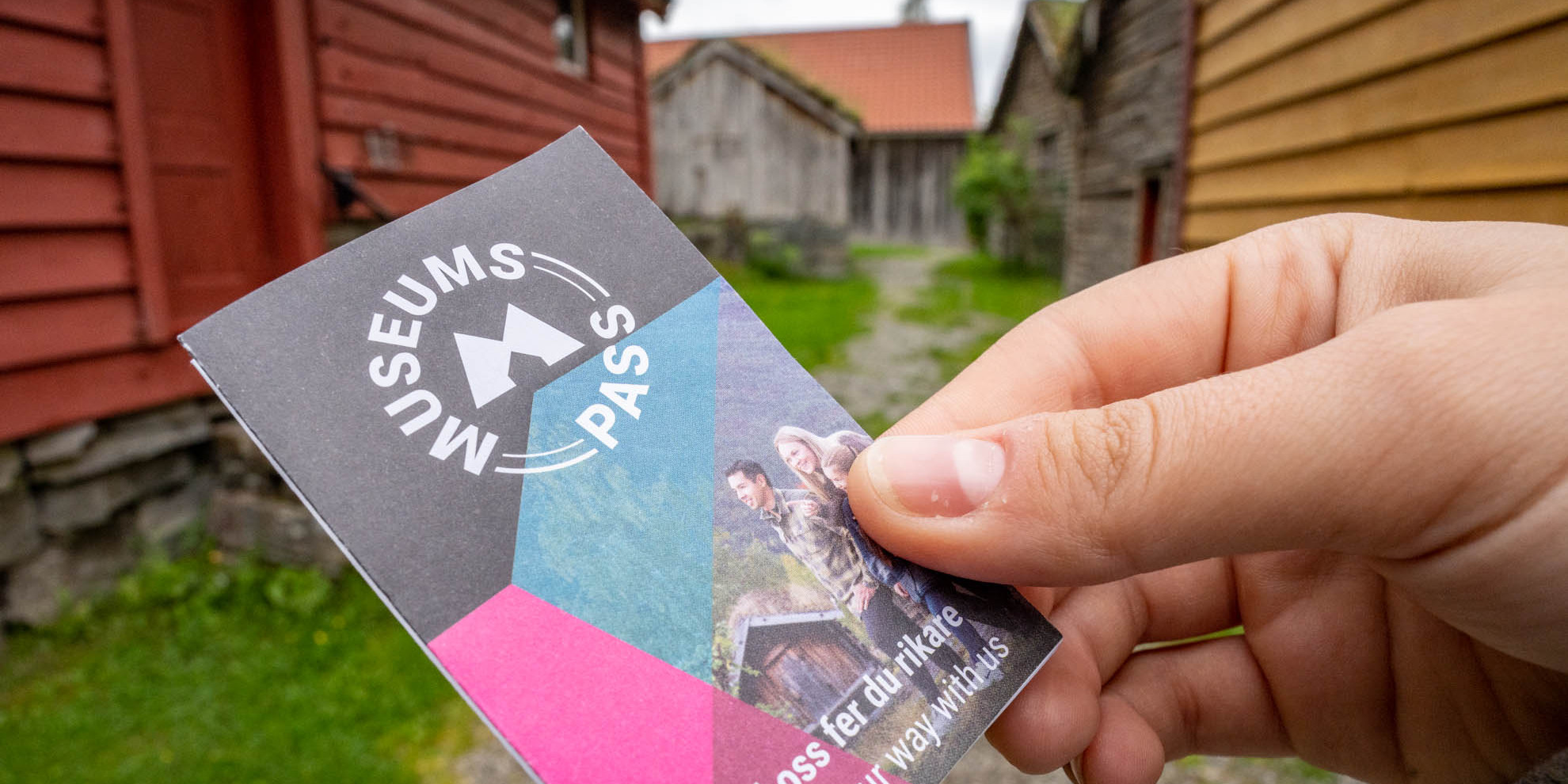 Ei hand held museumspasset mellom fingrane framfor museumsbygg.