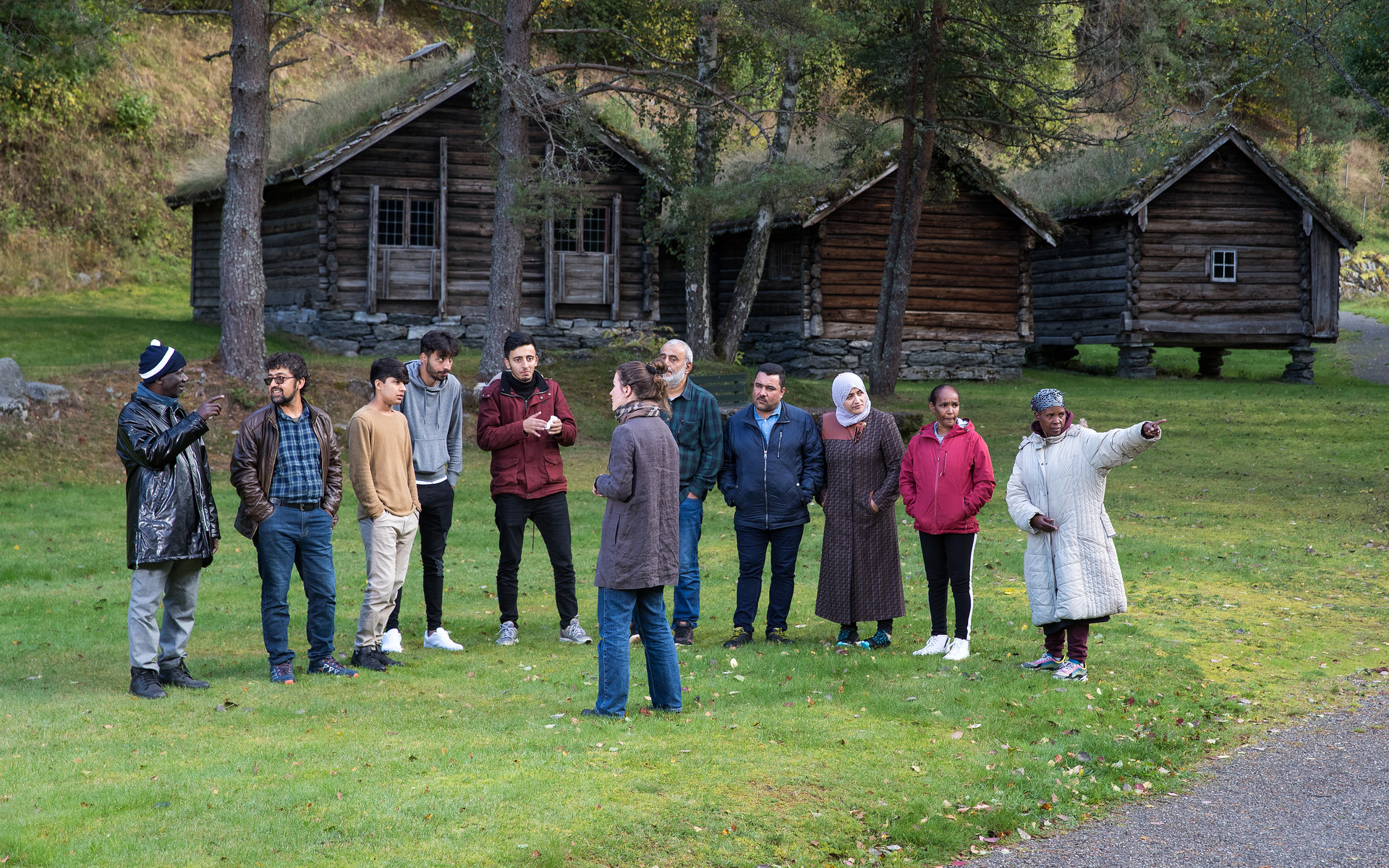 Guide med ei gruppe mennesker på omvisning i friluftsmuseet. I bakgrunnen ser vi hus frå 1800-talet, med gresstak og lafta veggar.