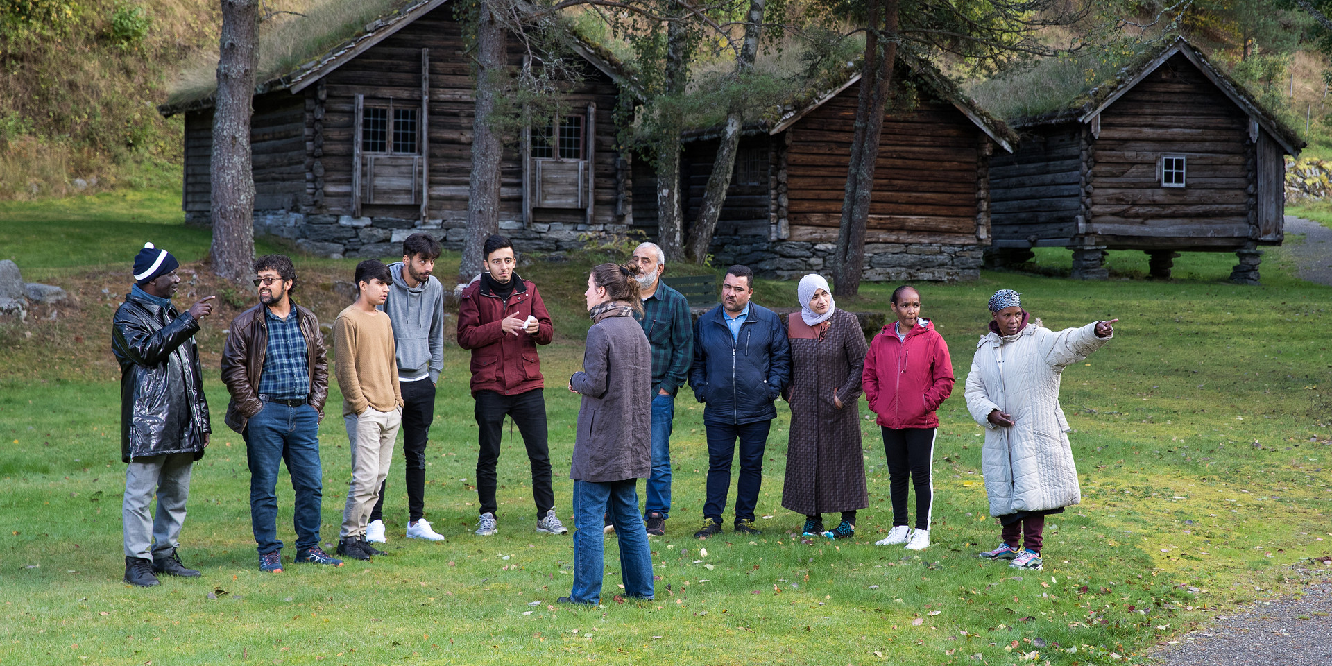 Guide med ei gruppe mennesker på omvisning i friluftsmuseet. I bakgrunnen ser vi hus frå 1800-talet, med gresstak og lafta veggar.