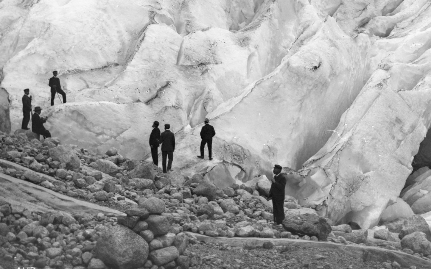 Svartkvitt-bilde av sju menn som står og studerar ein isbre.