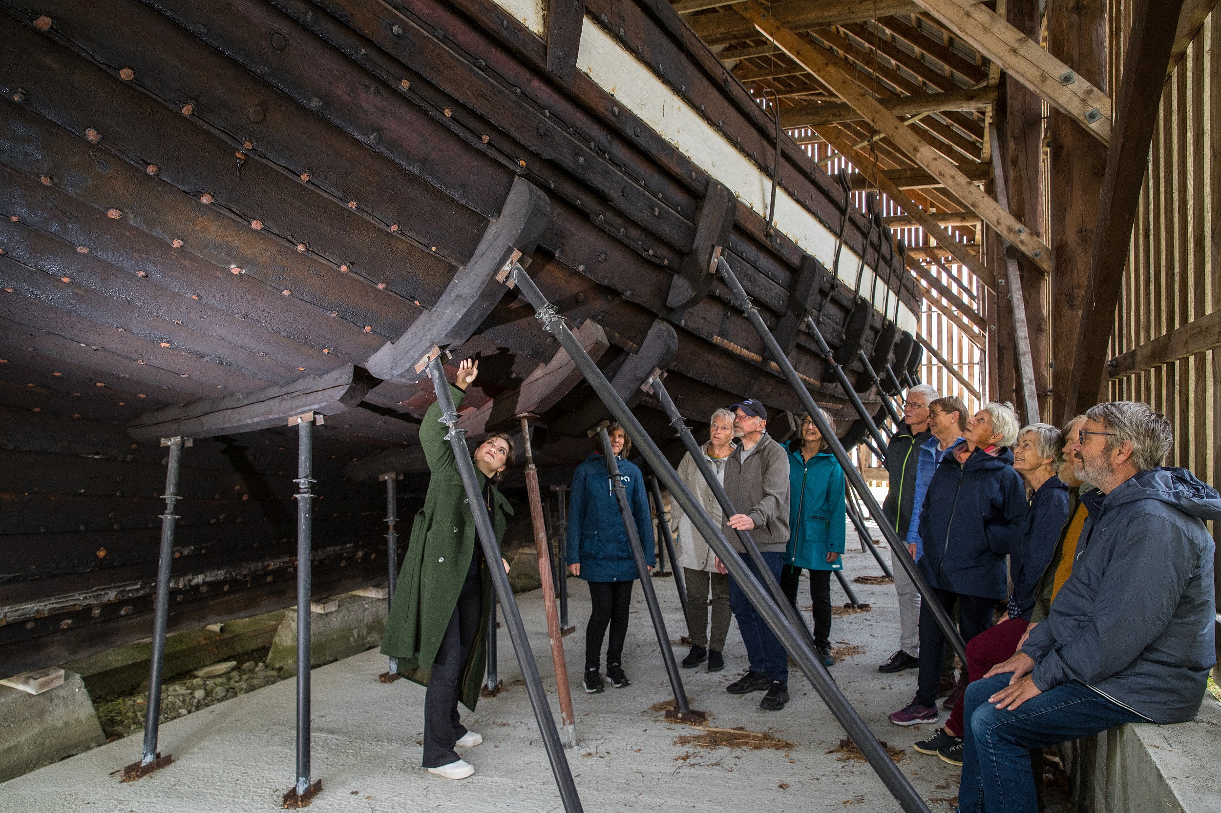 Gruppe med vaksne folk med guide på omvisning på Holvikejekta. Menneska står inne ved støttene til båten, og ser på undersida av jekta.