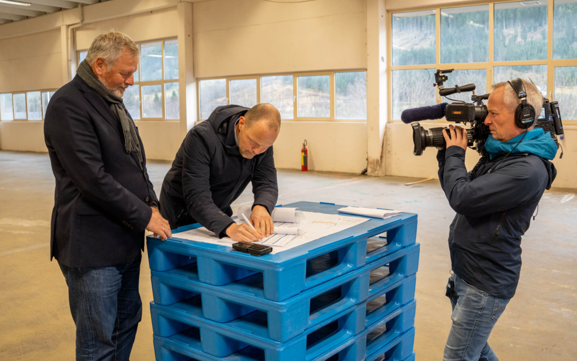 Signerer avtale som ligg på eit blått bord, kameramann frå NRK filmar.