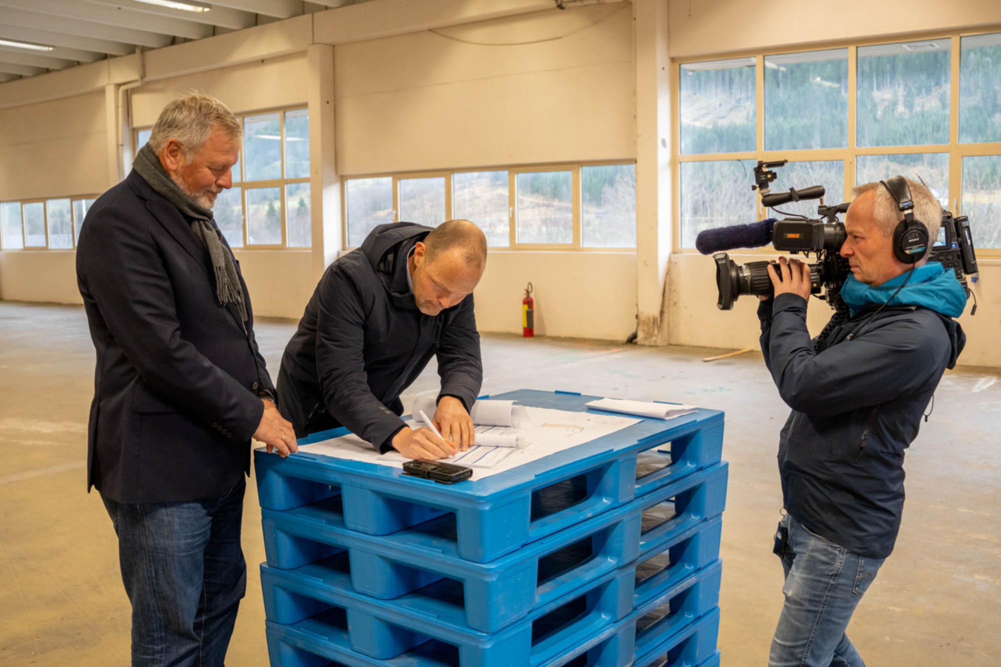 Signerer avtale som ligg på eit blått bord, kameramann frå NRK filmar.