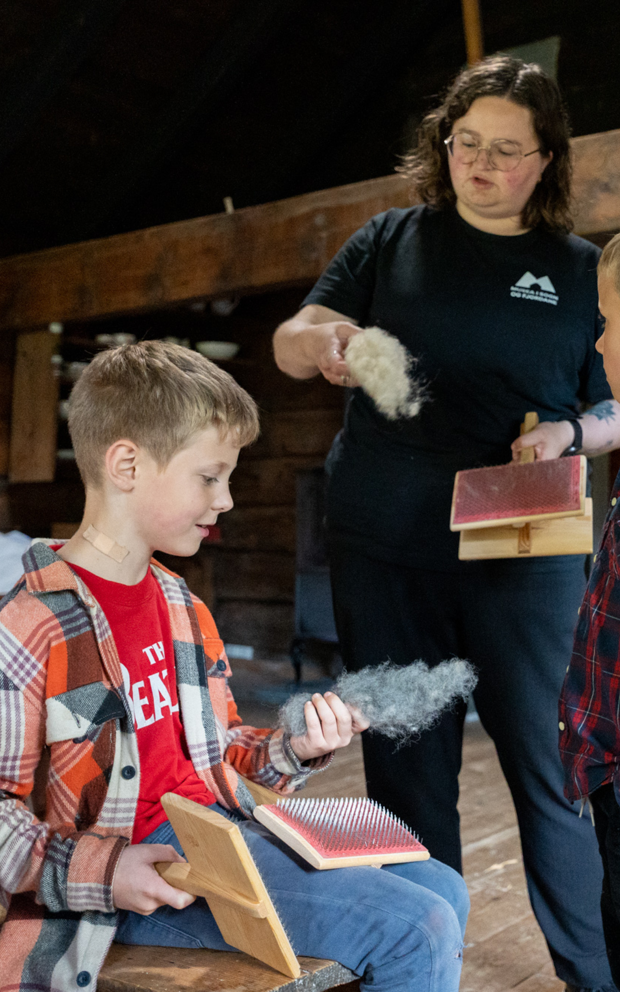 Kvinne viser to gutar korleis karde ull.