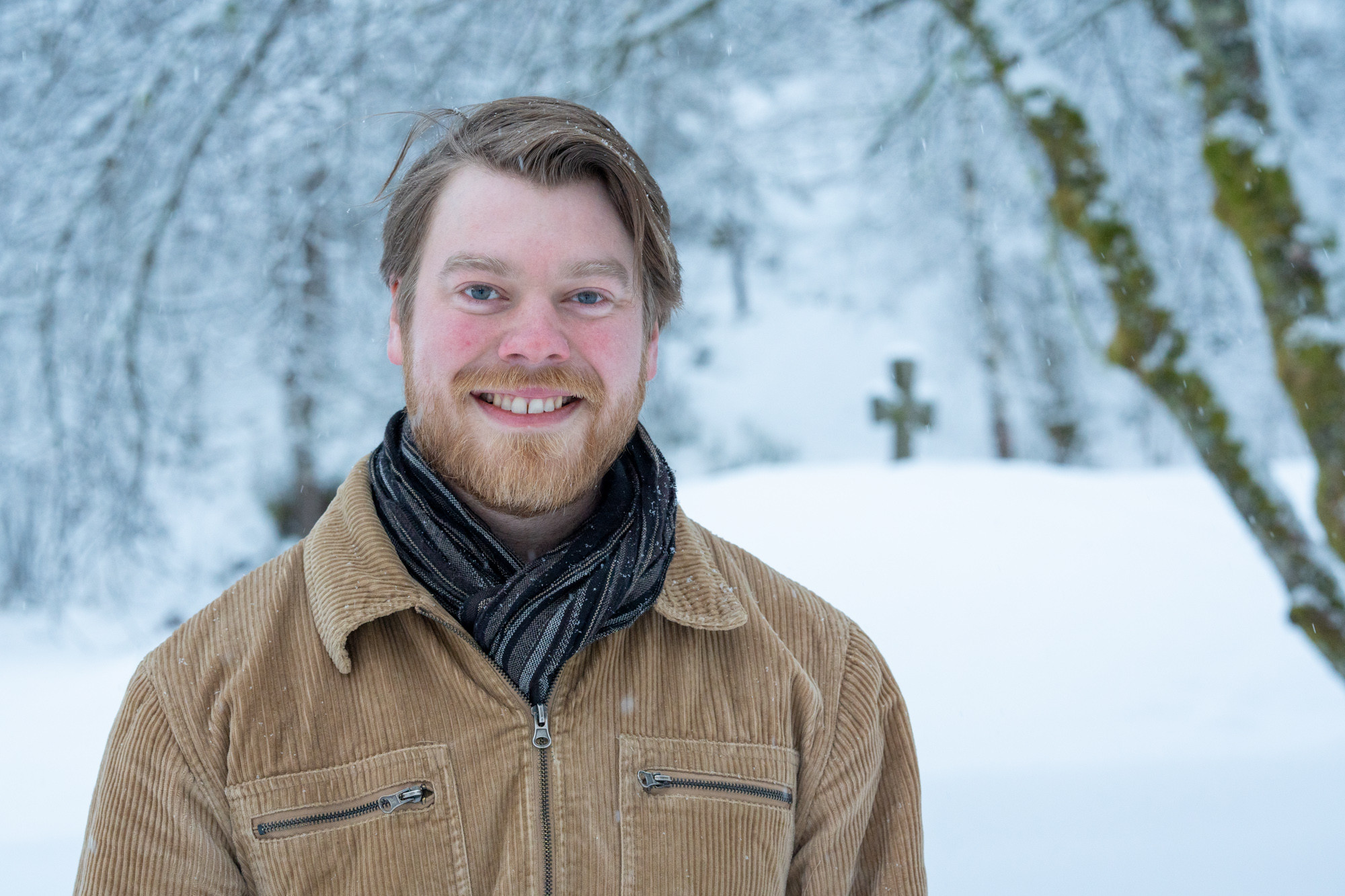 Portrettfoto av smilande mann med skjegg. Set er snø og i bakgrunnen skimtast ein steinkors.