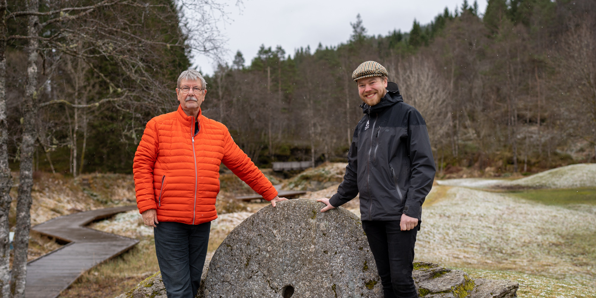 To menn støttar seg til ein ståande kvernstein, skog i bakgrunnen.