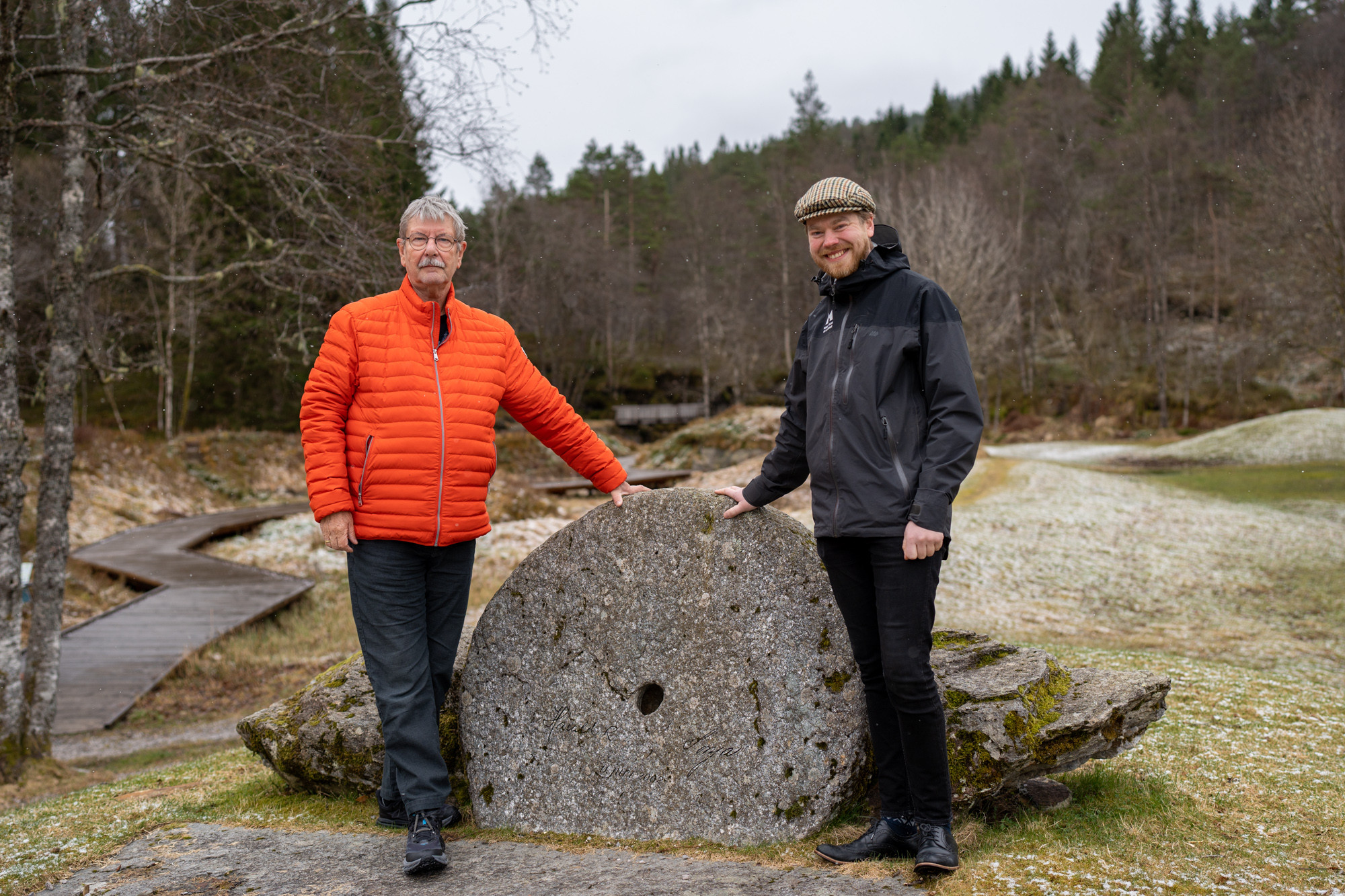To menn støttar seg til ein ståande kvernstein, skog i bakgrunnen.