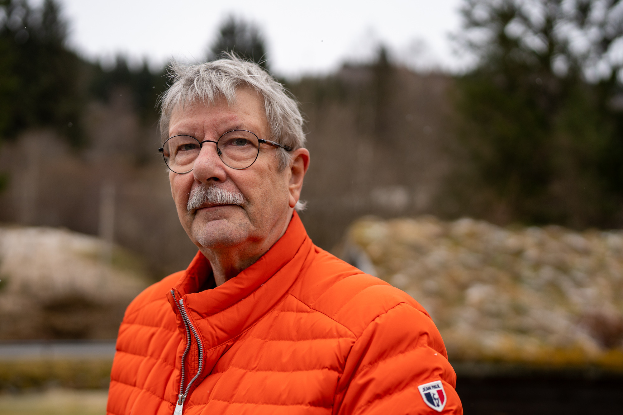 Portrettbilde av man i oransje boblejakke med gråleg hår, bart og briller med nøytralt uttrykk.