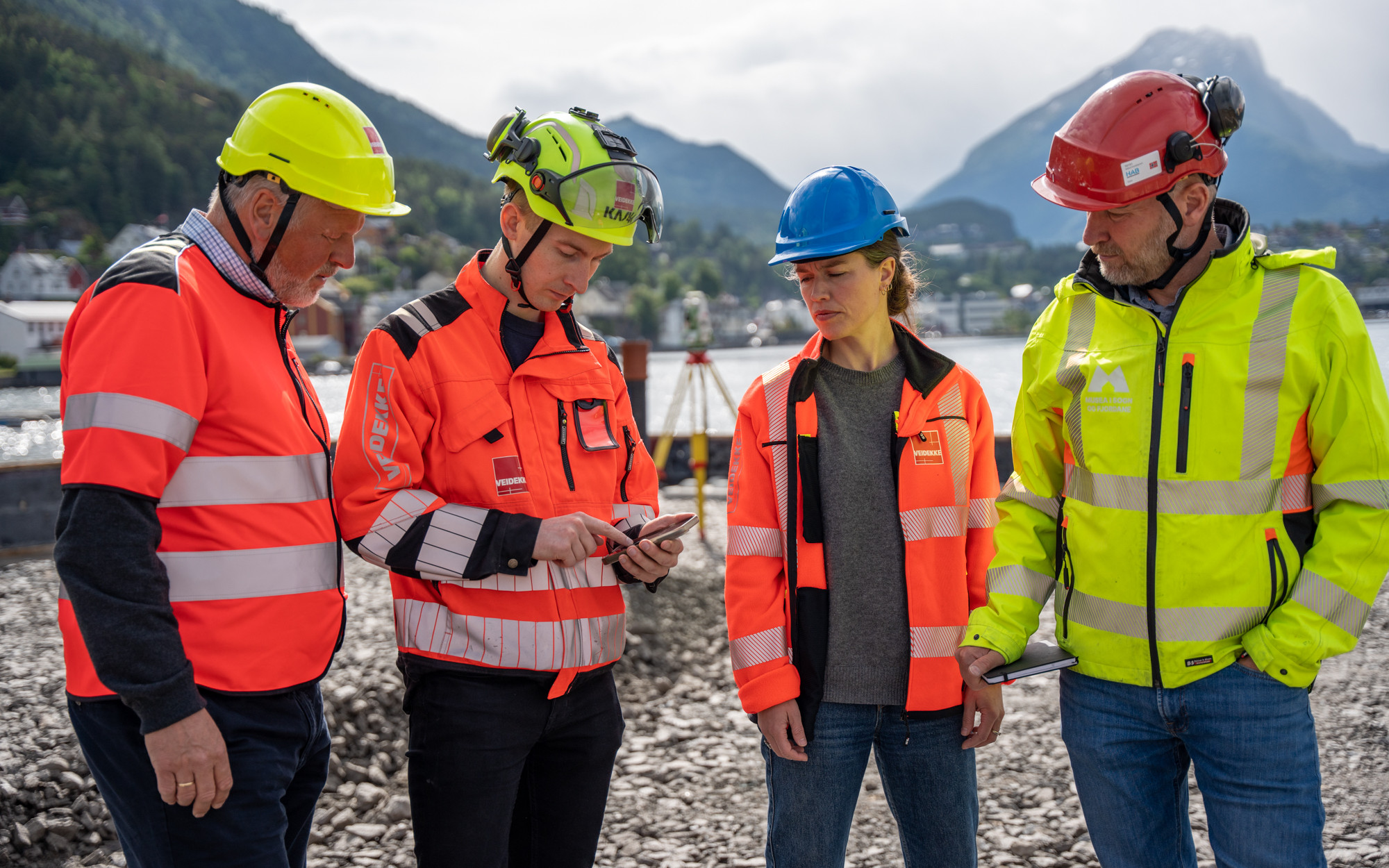 Fire personar med hjelm og signalfarga vestar står på ein byggeplass med fjell i bakgrunnen.