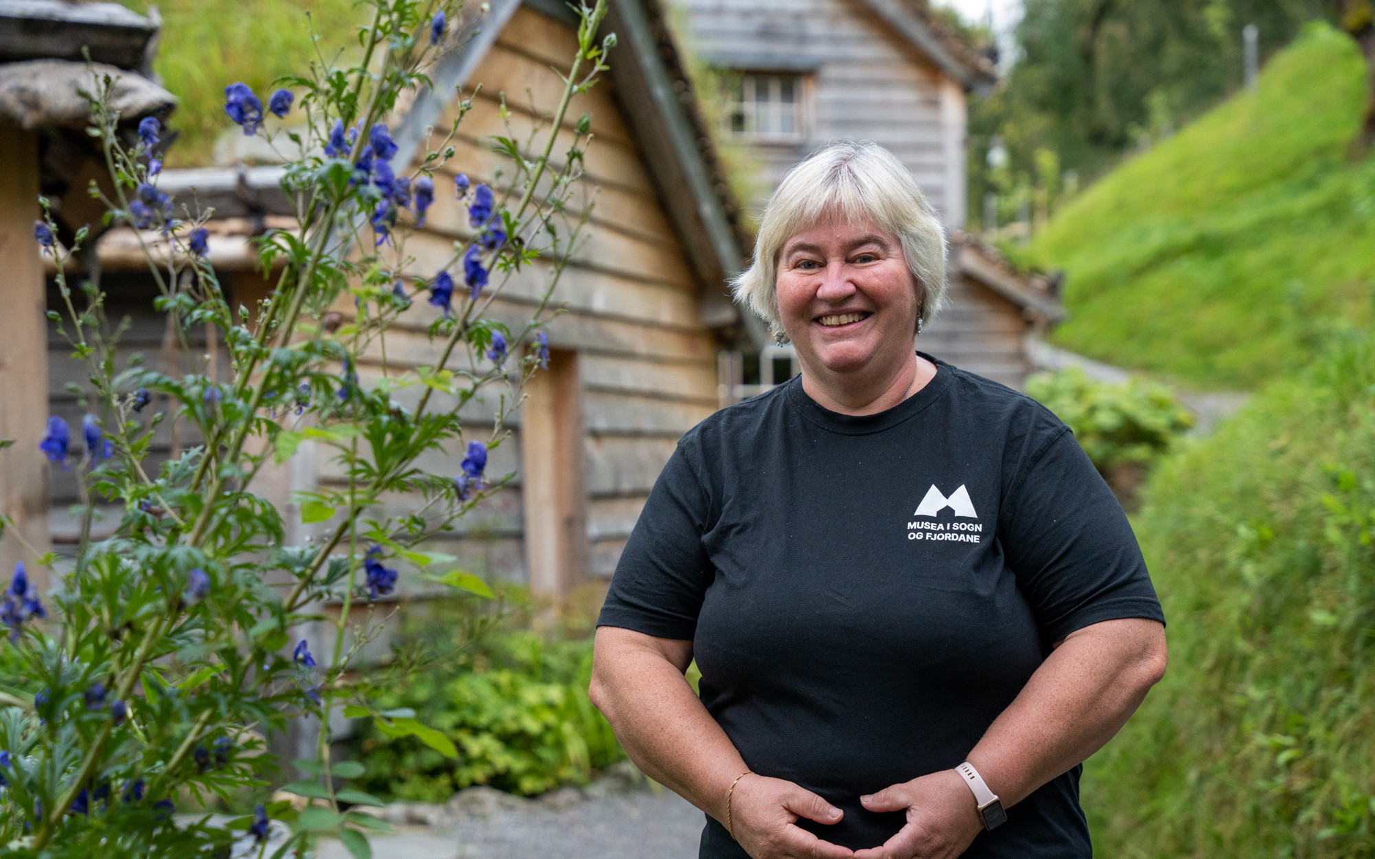 Smilande kvinne med grått hår og svart t-skjorte framfor gamle trebygningar med torvtak