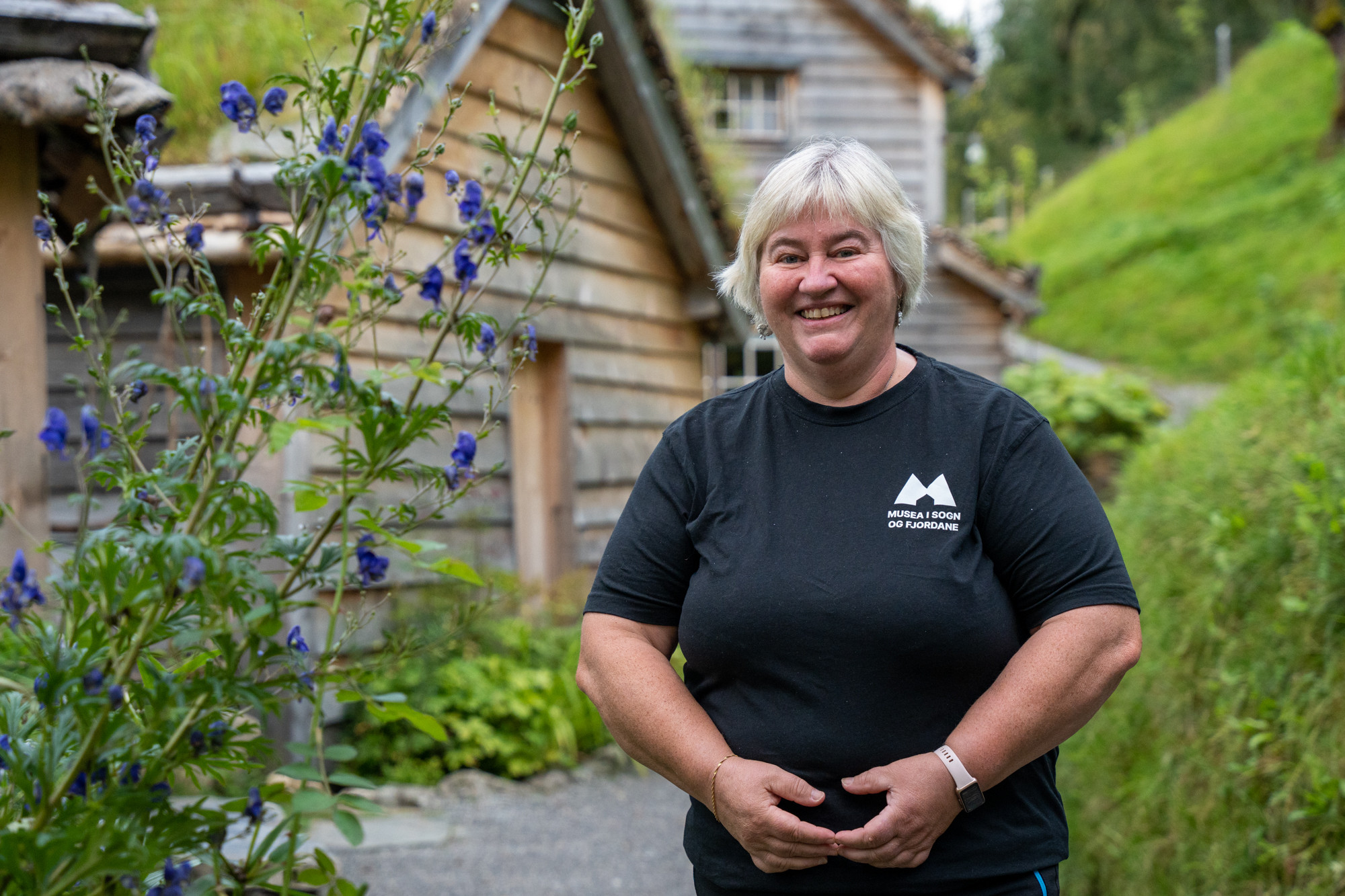 Smilande kvinne med grått hår og svart t-skjorte framfor gamle trebygningar med torvtak.