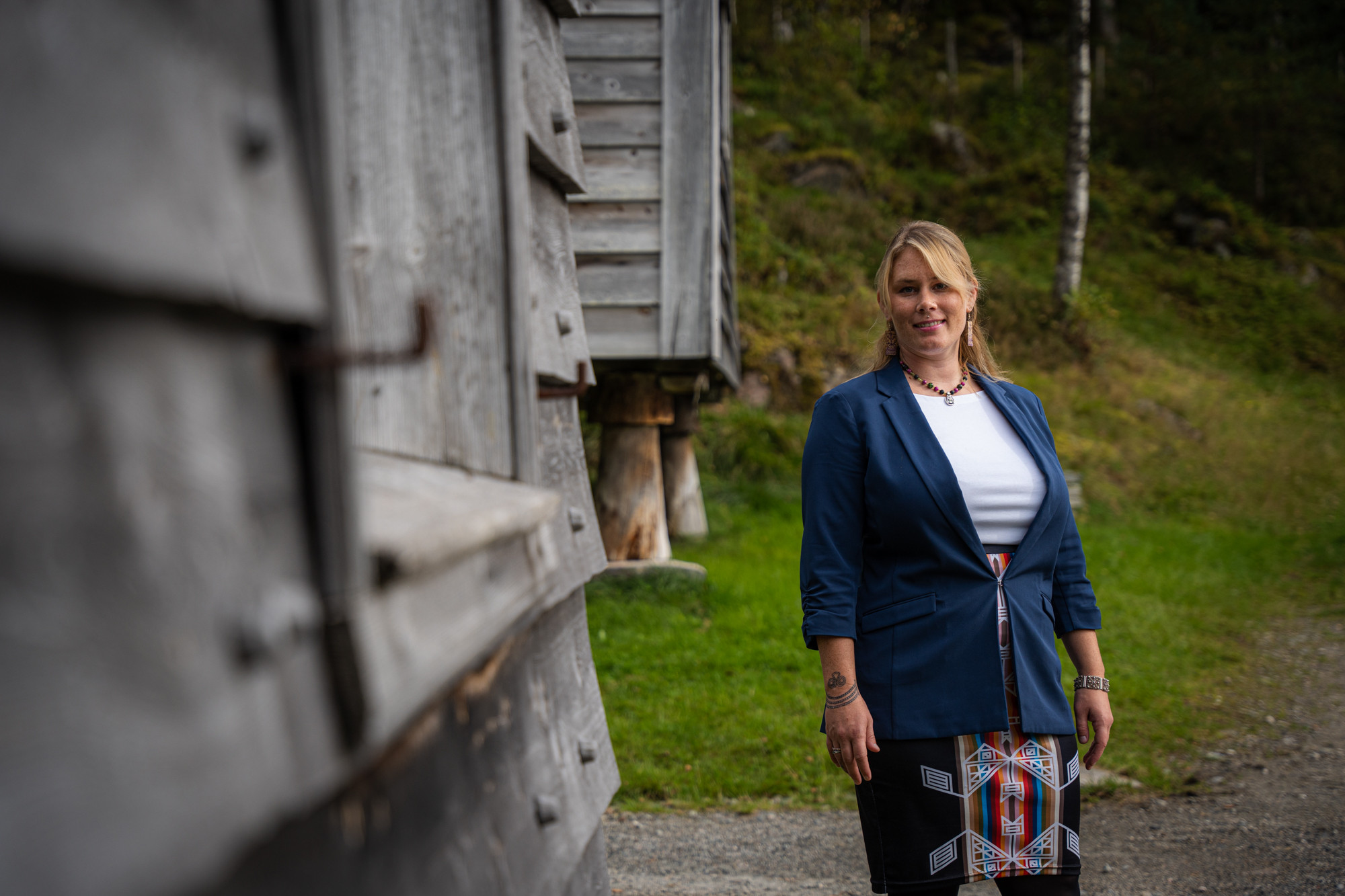 Ung lyshåra kvinne med blå dressjakke framfor gamal trebygning.