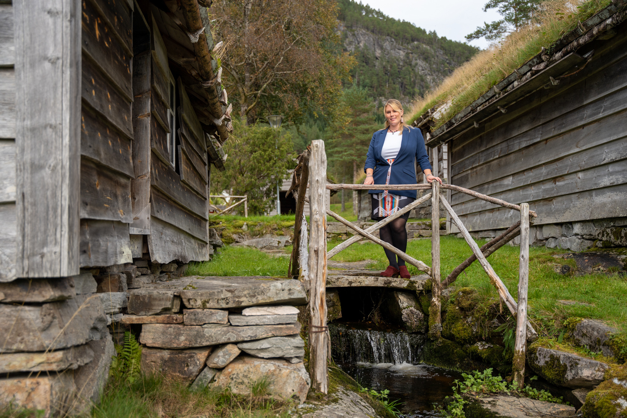 Kvinne står bak rekkverk på steinhelle over liten bekk mellom tømmerhus.