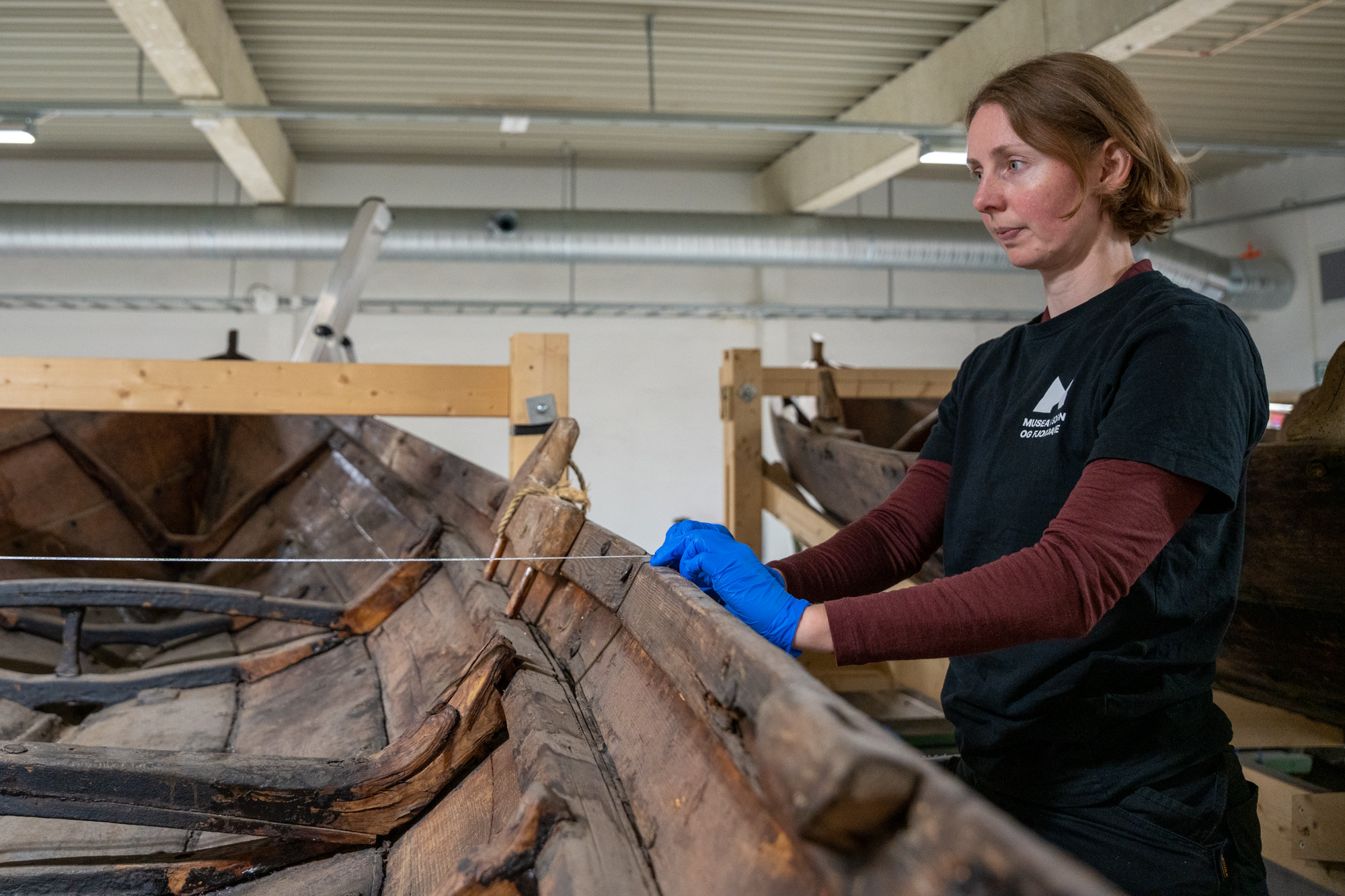 Kvinne med blå eingongshanskar held eit måleband over ripa på ein gamal trebåt.