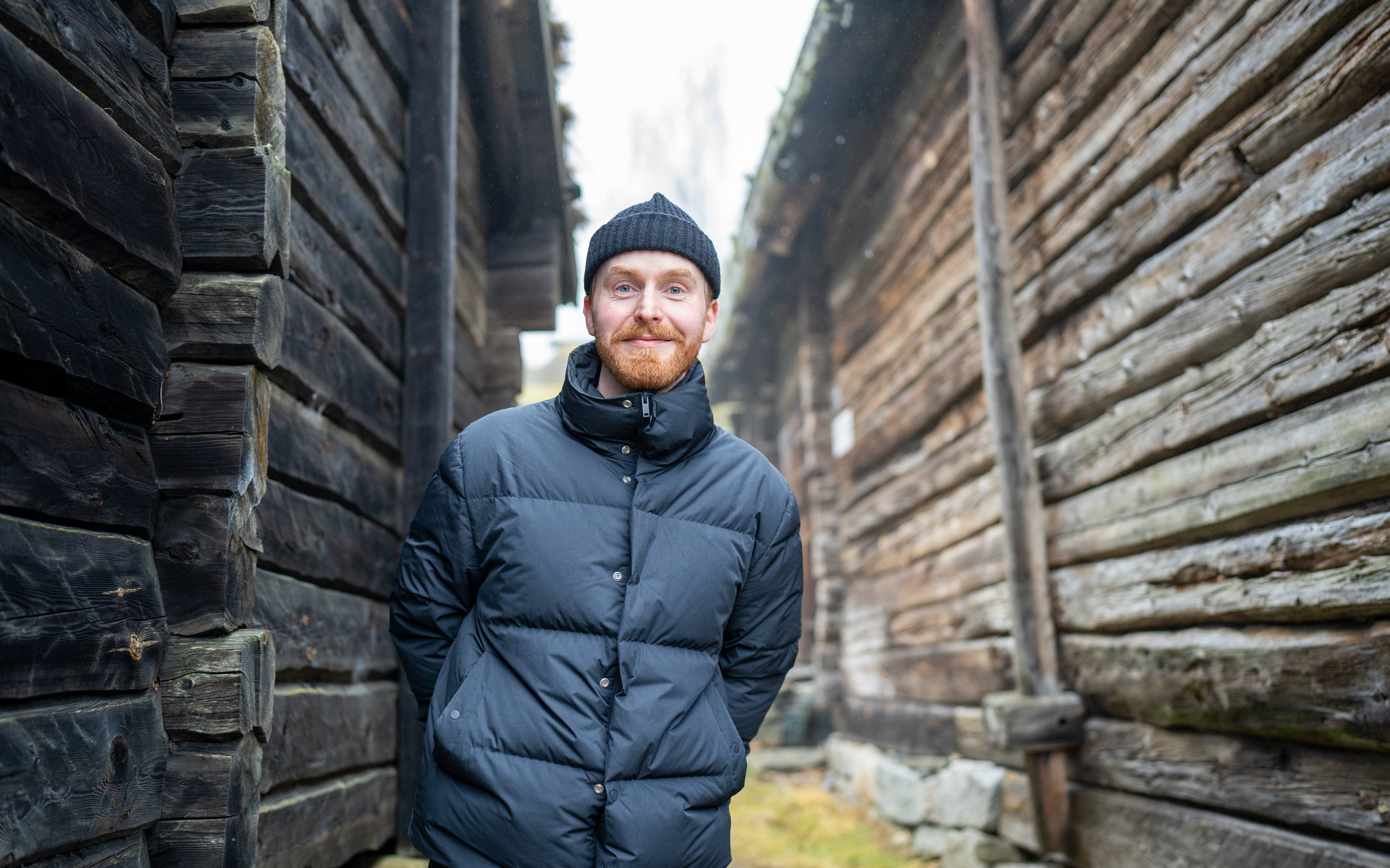 Mann med skjegg mellom to gamle lafta tømmerhus