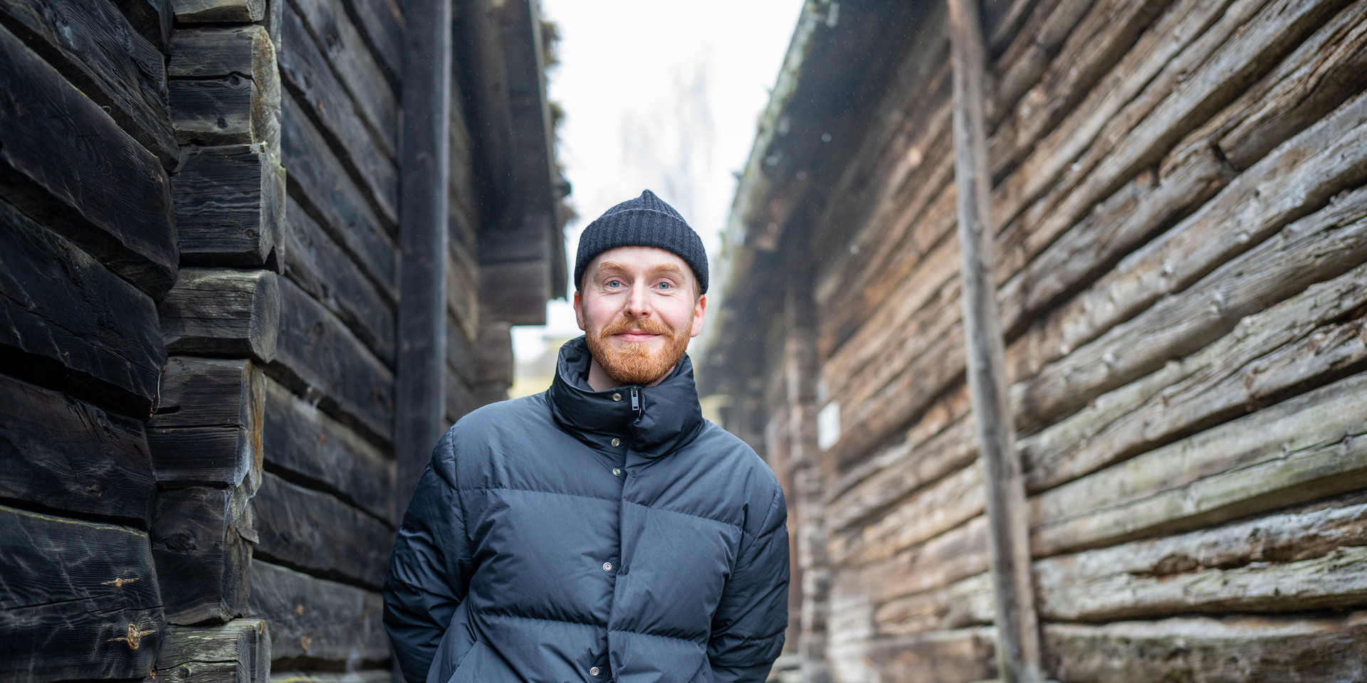Mann med skjegg mellom to gamle lafta tømmerhus.