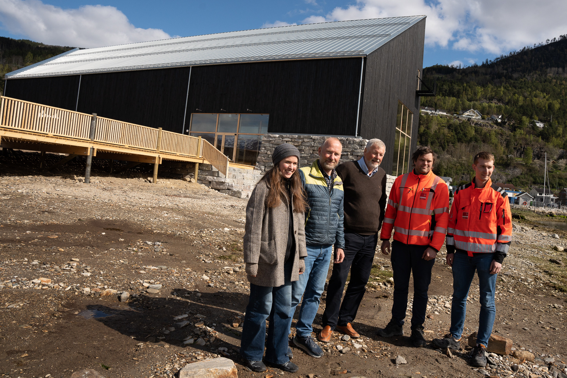 Fem personar står i strandkanten med svart, nytt bygg i bakgrunnen.