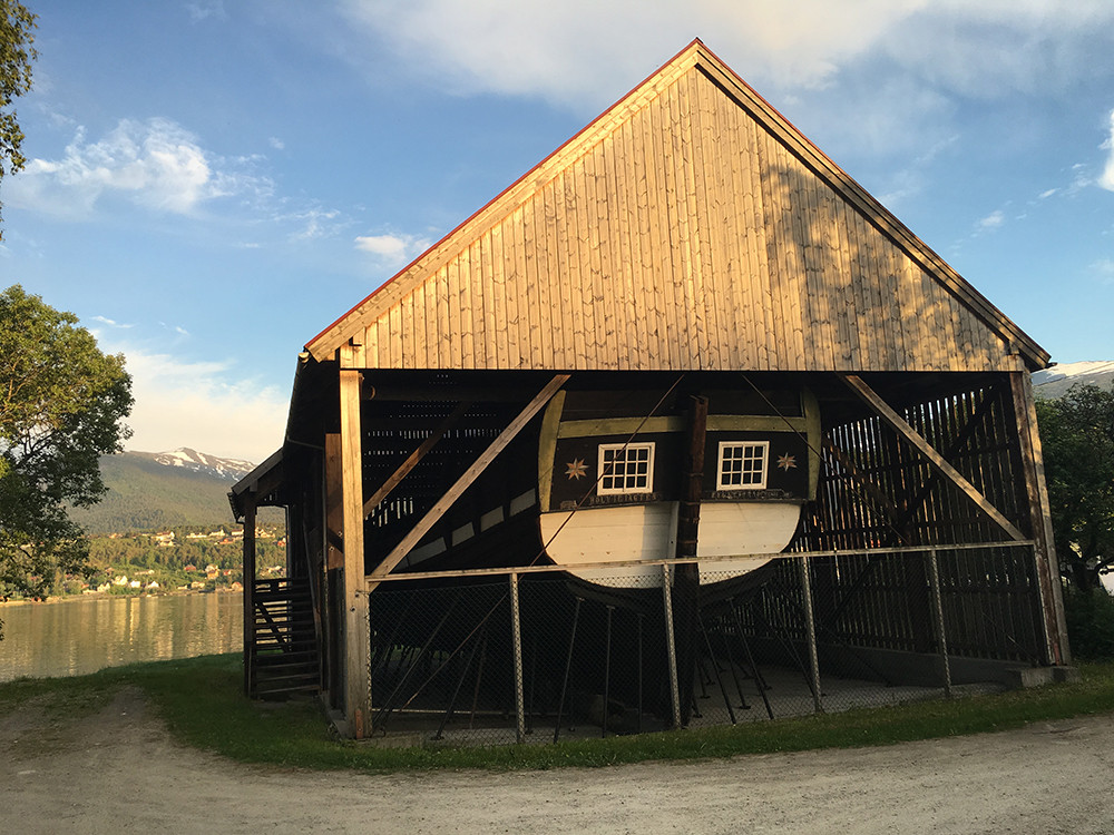 Gamal trebåt under eit tak med kvitmåla, avkutta akterstamn. Under ein blå himmel i solnedgang ved fjorden.