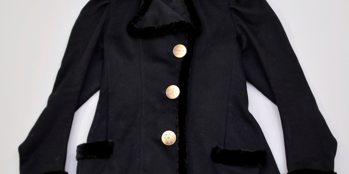 Innsvingt, svart jakke. Alle kantar er kanta med flossa stoff. Opning framme, litt på sida. Knapping med knapphol. Store, knappar med dekor.