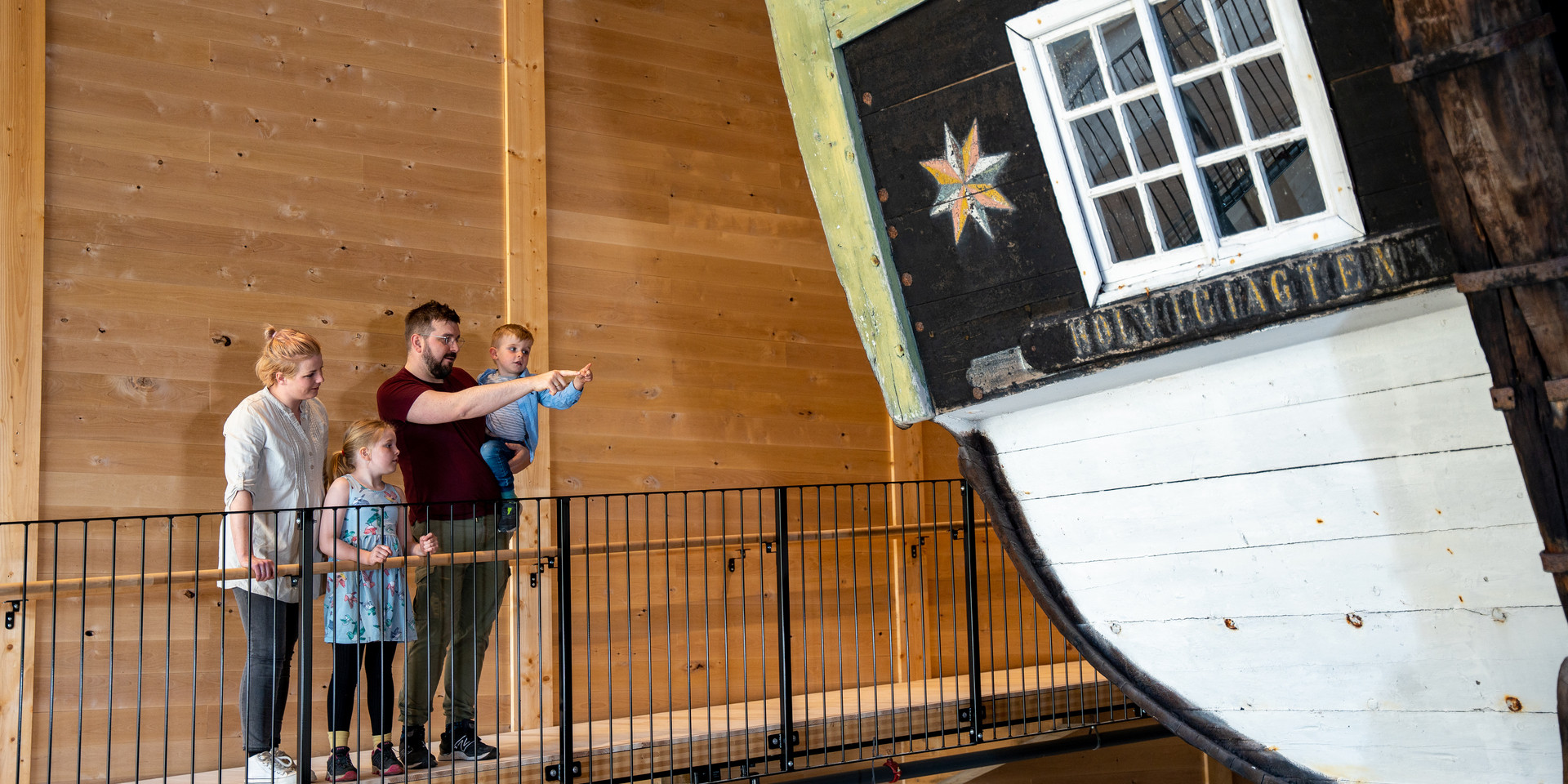 To born og to vaksne står og ser og peikebr på ein stor båt inne i eit museum frå sida.