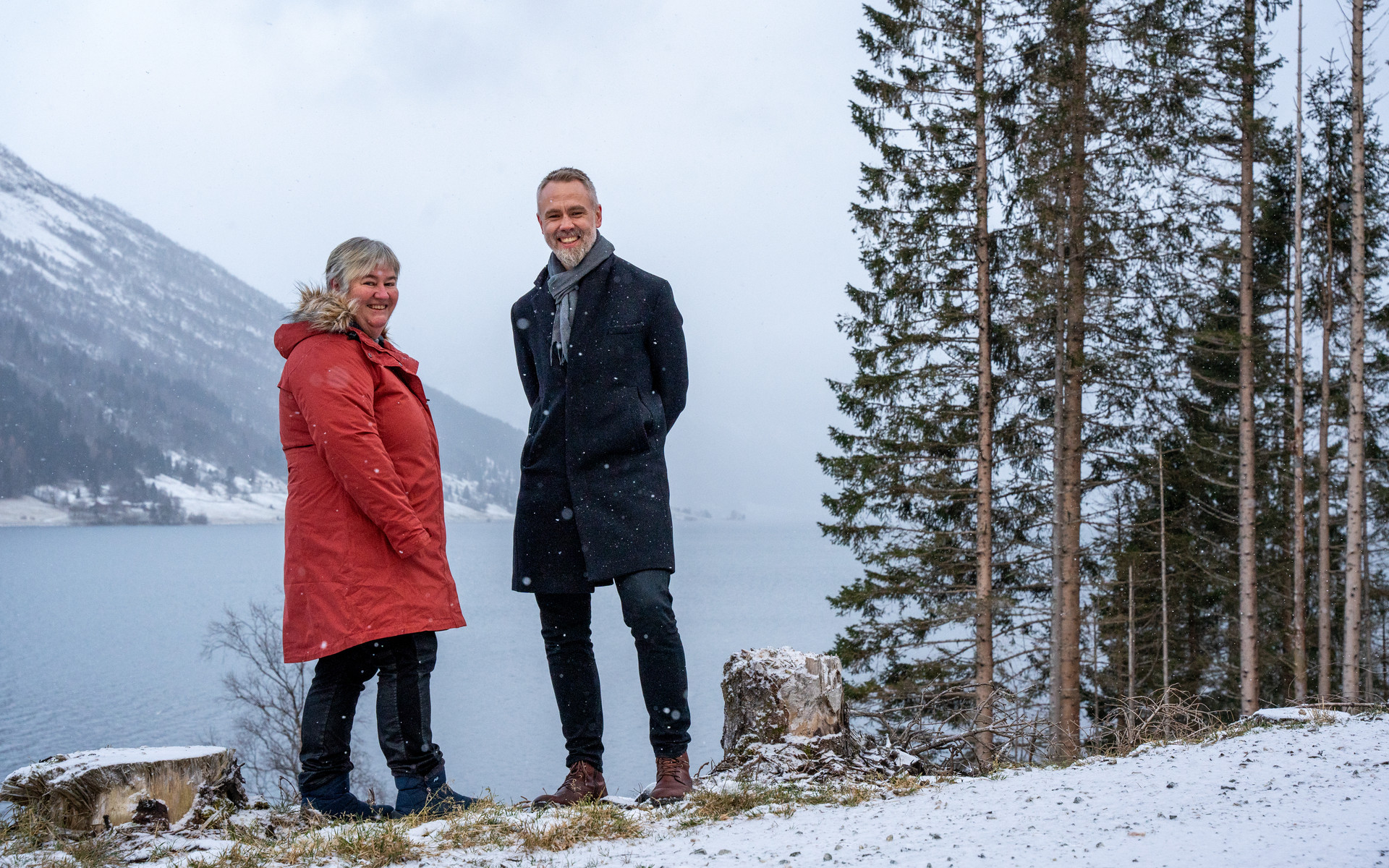 Ein smilande mann og kvinne i vinterkle og lett snøver. Omkransa av stubbar, graner og jølstravatnet og eit fjell i bakgrunnen.