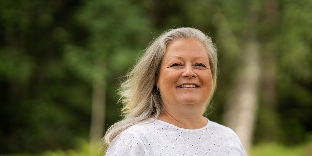 Smilande kvinne med kvit skjorte med tre i bakgrunnen