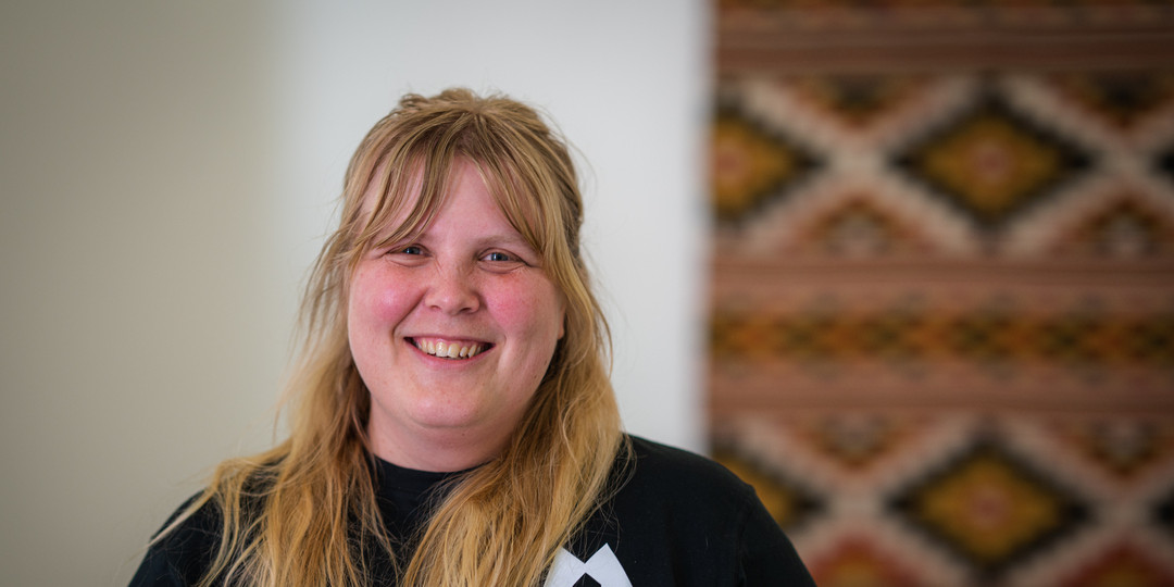 Portrett av smilande kvinne framfor ein vevd duk på vegg