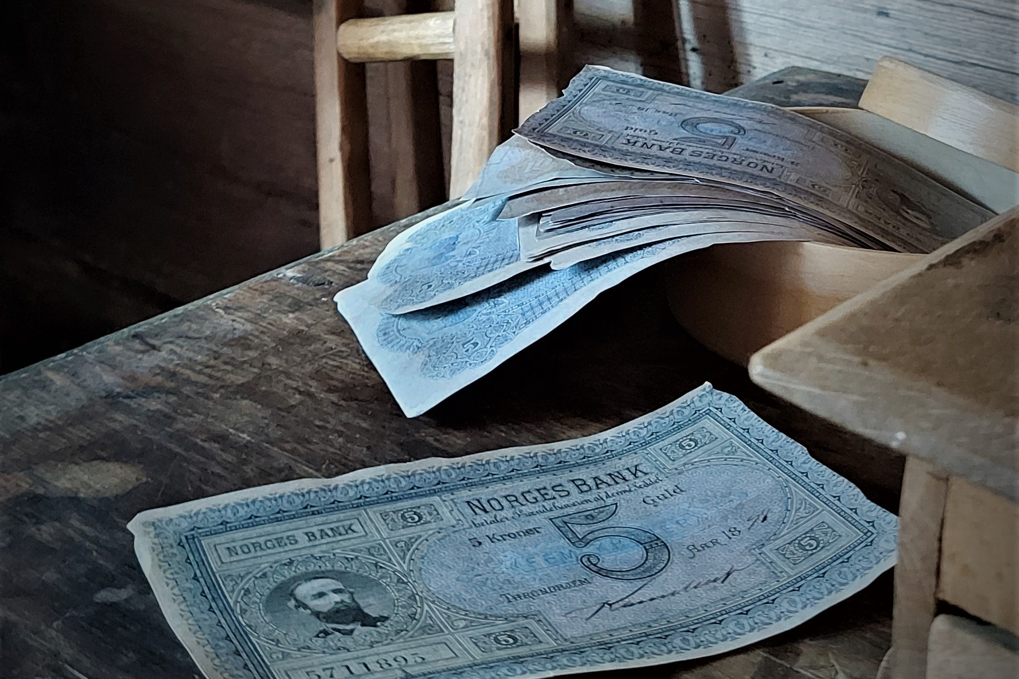 Eit skrin med gamle pengesetlar på eit trebord i eit lafta hus. Bak skrinet står eit par gamle krykker i tre inntil ein vegg og eit skåp.