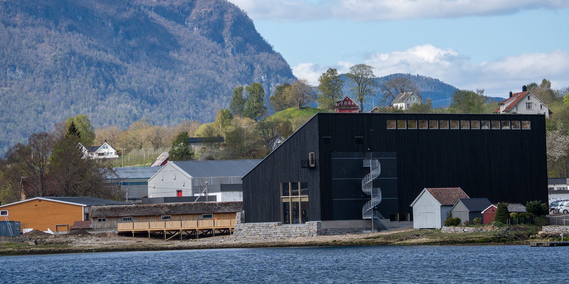 Bilde av et stort sort bygg. På fremsiden er der en lang vindeltrapp som er festet i bygget. Til høyre for bygget ser vi noen små naust. I bakgrunnen av bygget ser vi trær, fjell og hus. Fremfor bygget ser vi vann.