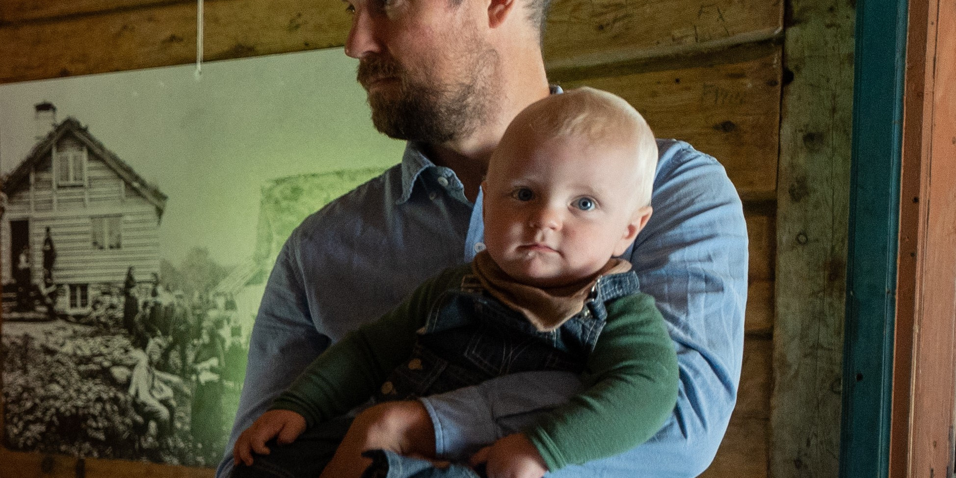Far og barn i gammelt hus. Historisk fotografi i bakgrunnen.
