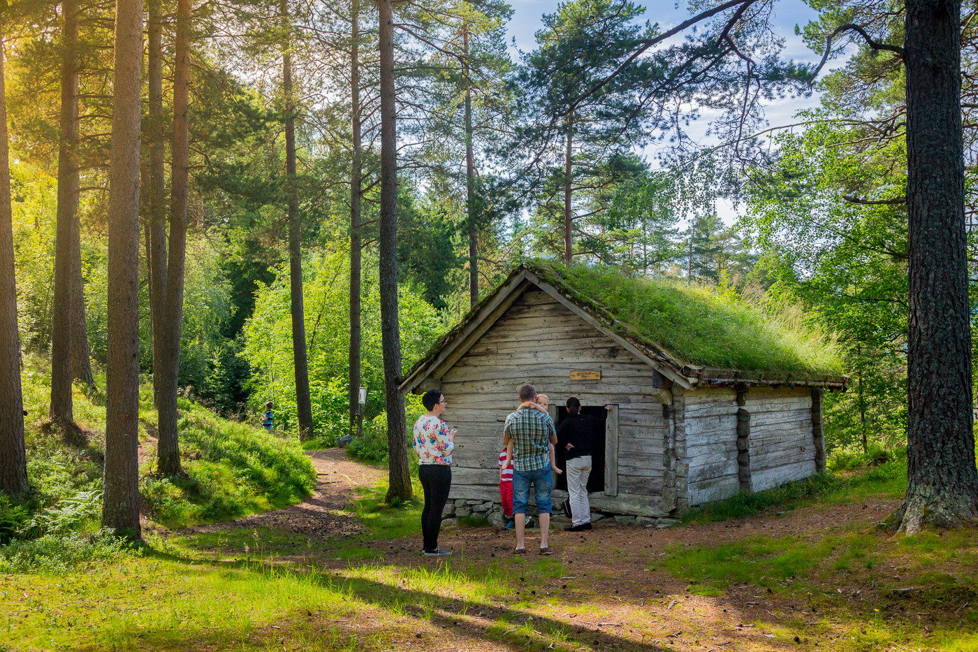 Familie på veg inn i eit støylshus i eit skogsområde. Huset har torvtak og tømmerveggar i lafta stil.
