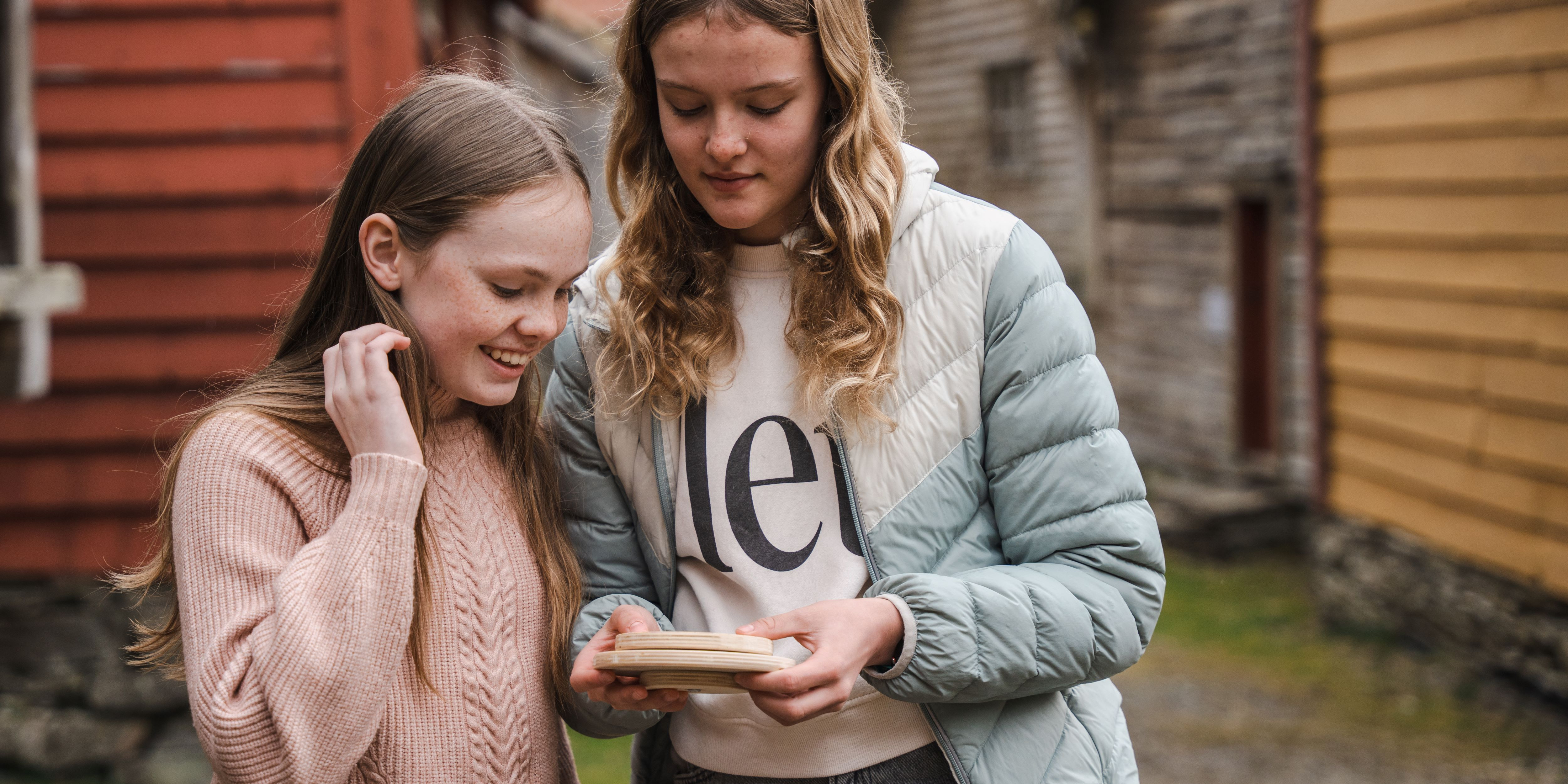 To unge jenter som holder på et hjul. I bakgrunnen ser vi et rødt hus, et gult hus og tre brune hus.