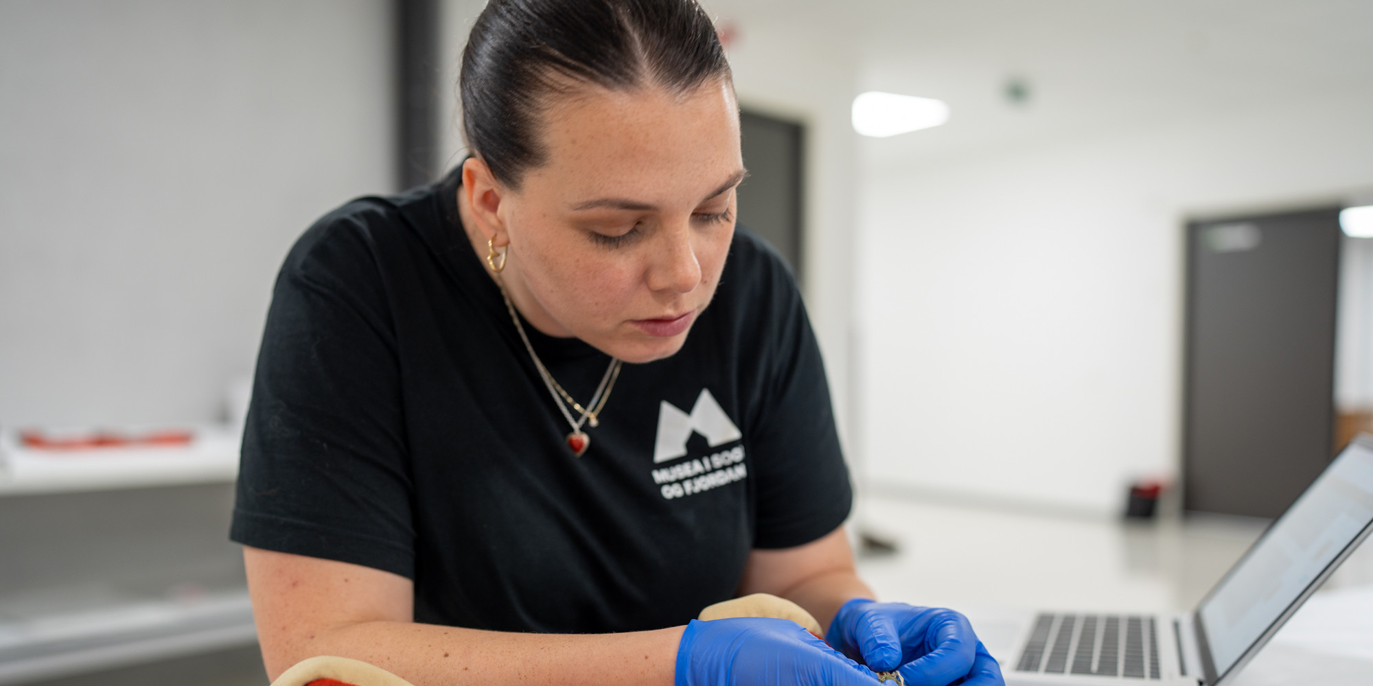 Kvinne med blå latex-hanskar undersøker eit raudt folkedraktsliv.
