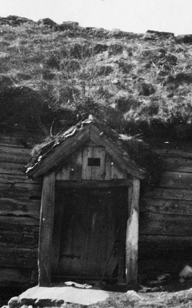 Svart-kvitt bilde av eit skakt hus med torvtak.