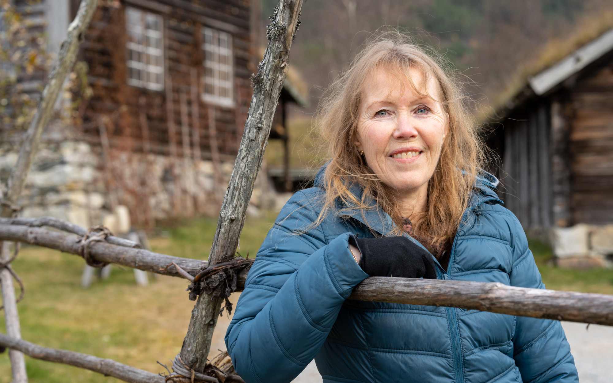 Aud Ross Solberg med langt gyllent hår og blå boblejakke lener seg på ei hesje framfor gamle museumsbygningar.