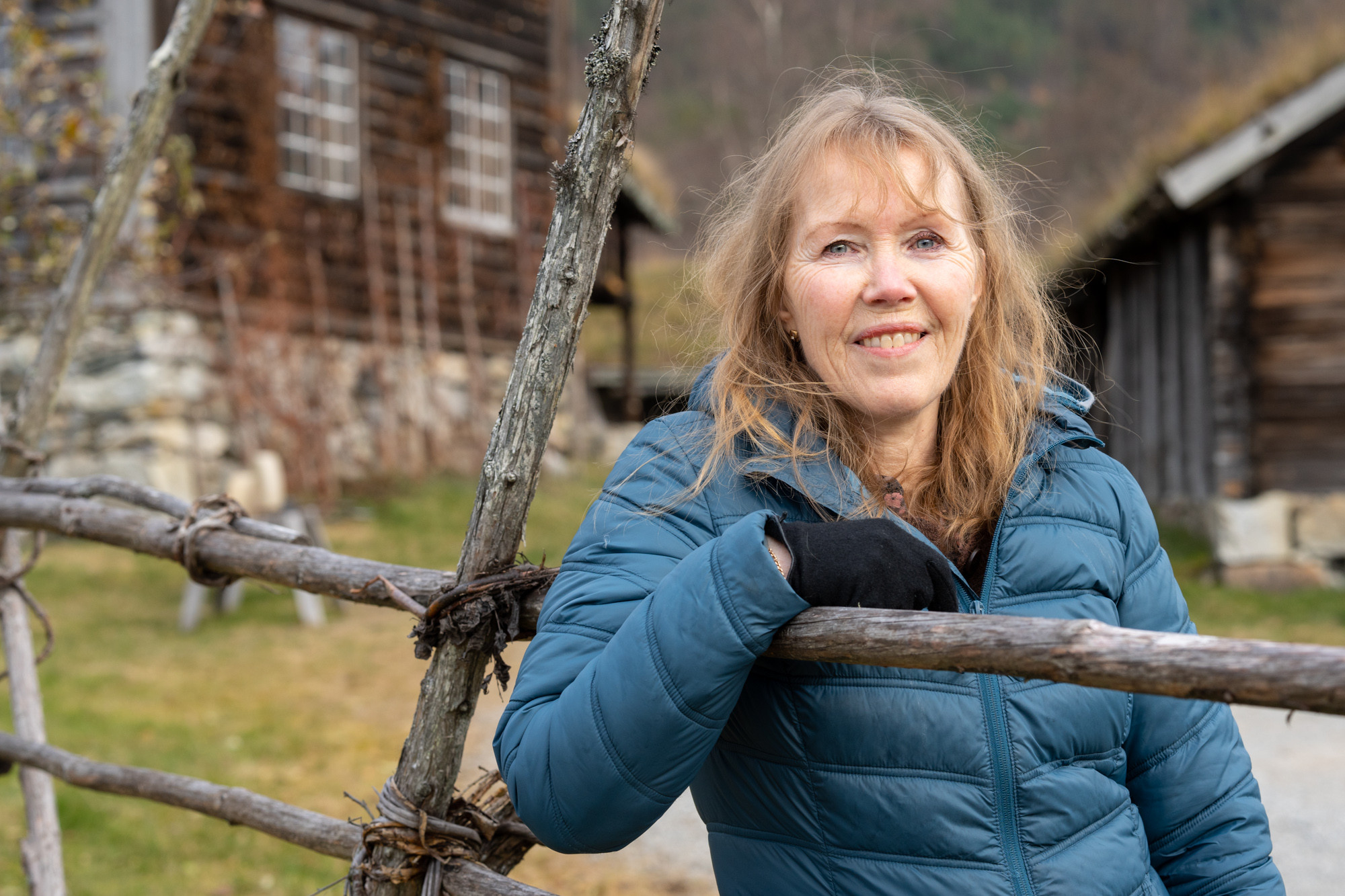 Aud Ross Solberg med langt gyllent hår og blå boblejakke lener seg på ei hesje framfor gamle museumsbygningar.