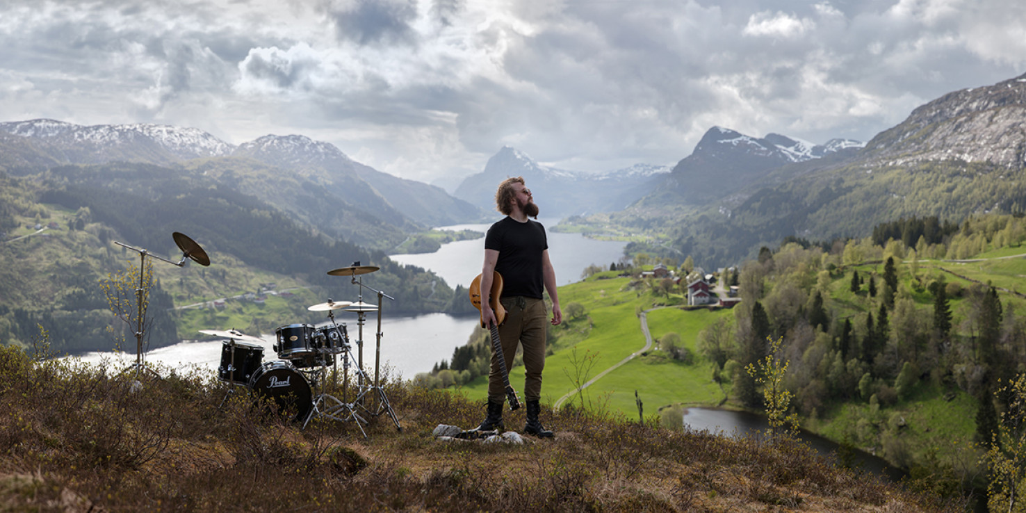 Verksfoto av fotoverket Gaular av Oddleiv Apneseth. I midten av bildet står en mann, som ser opp mot høyre. I hånda hans har han en trehvit el-gitar.  Til venstre i bildet, stående på bakken, er et trommesett. Bakgrunnen er storslått, med fjord og fjell. Himmelen viser stormskyer som kommer innover, men det er lysglimt ned i dalen.
