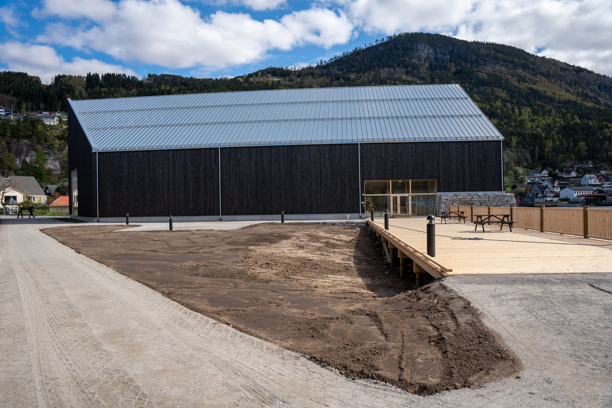 Svart bygg med grusveg, brygge og svart jord i forgrunnen.