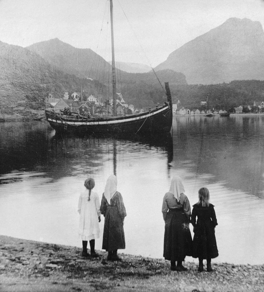 Fire kvinner i kjole og skaut står os er ut på fjorden, der ei jekt ligg ankra opp.