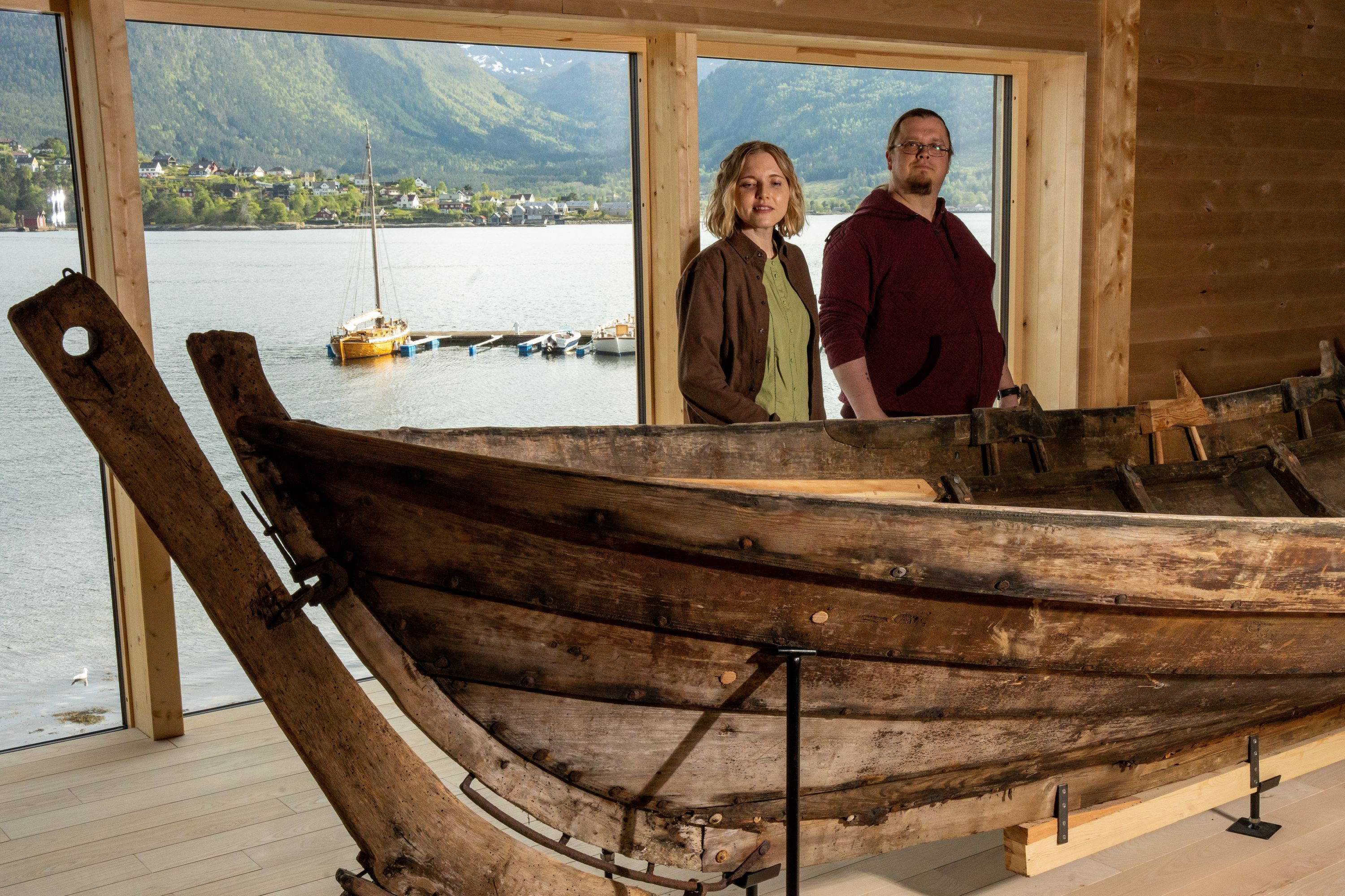 En kvinne og en mann ser på en robåt. I bakgrunnen ser man utsikt over fjord og natur.