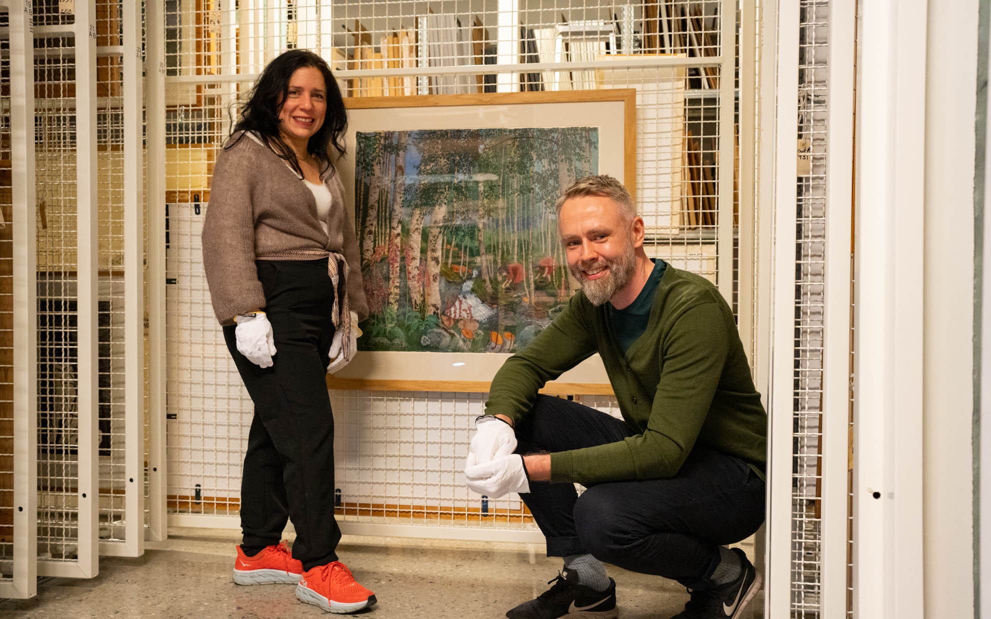 Mayra står og Tor Martin sit på huk framfor fargetresnittet "Revebjeller" som heng på ein kvit gittervegg i magasinet på Kunstmuseet.