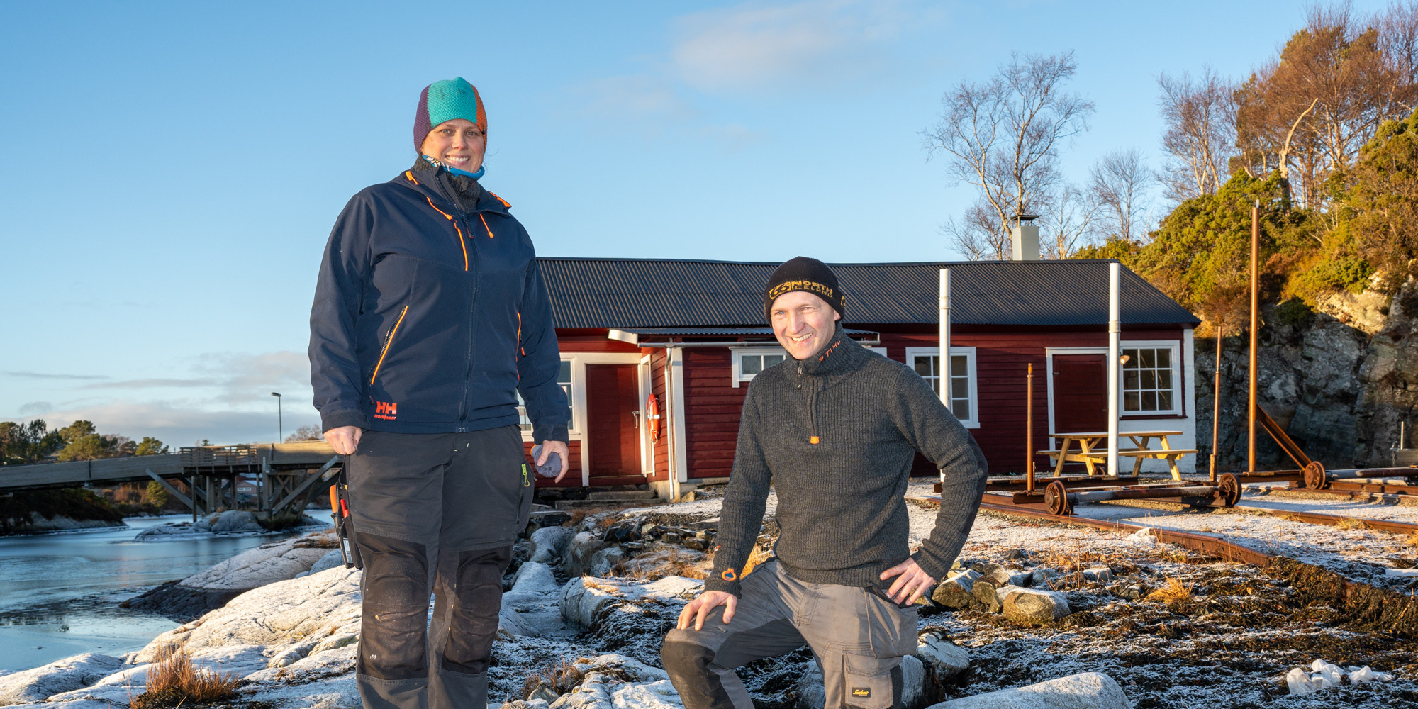 Mann og kvinne i arbeidstøy på eit svaberg, framofor eit lågt raudmåla hus med jernbanskinner framfor.