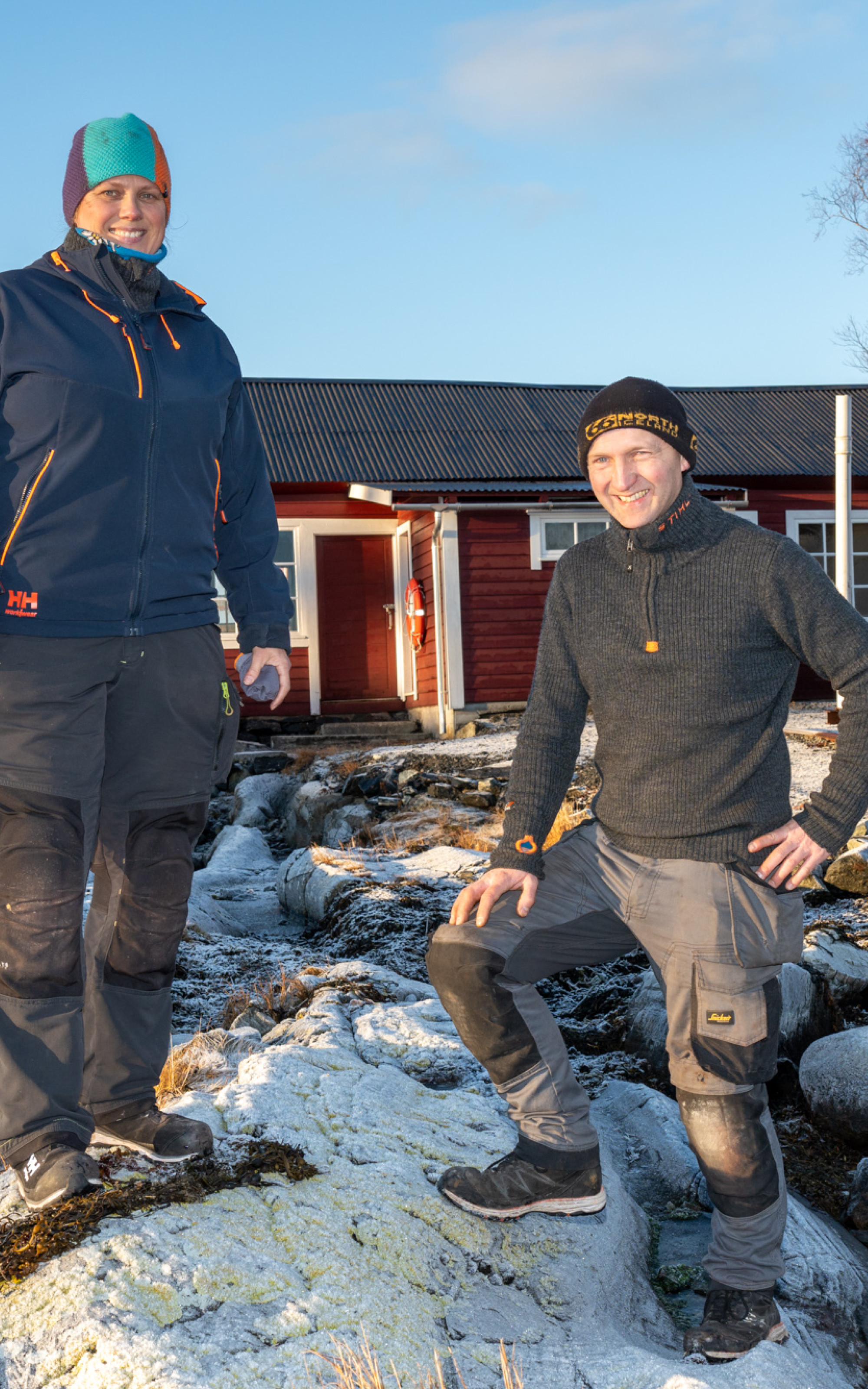 Mann og kvinne i arbeidstøy på eit svaberg, framofor eit lågt raudmåla hus med jernbanskinner framfor.