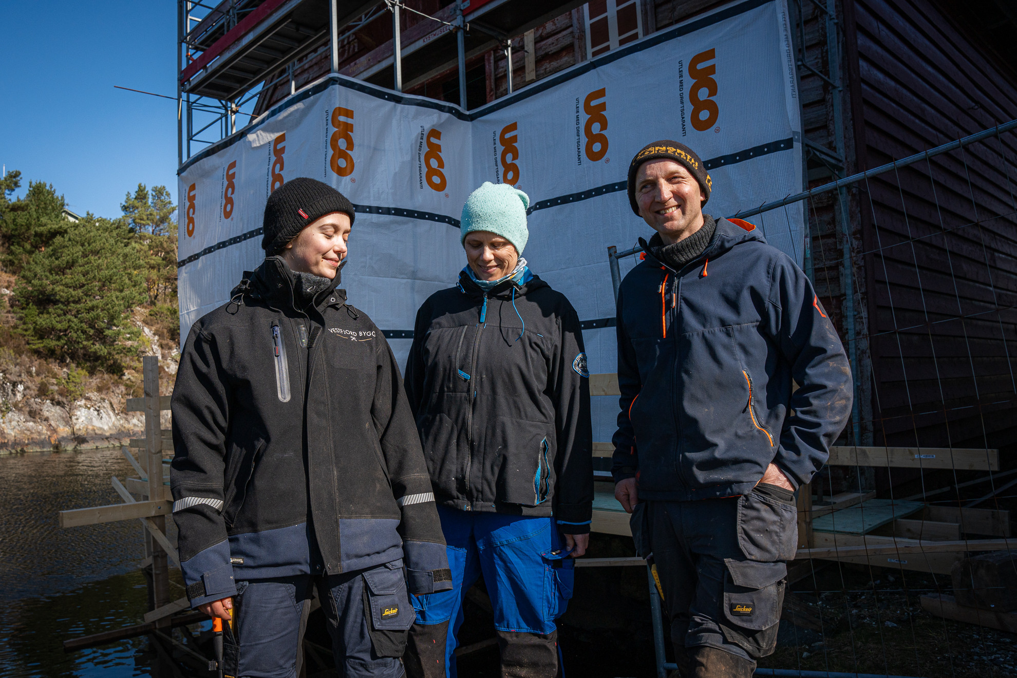 Tre personar i arbeidsklede framfor stillas.