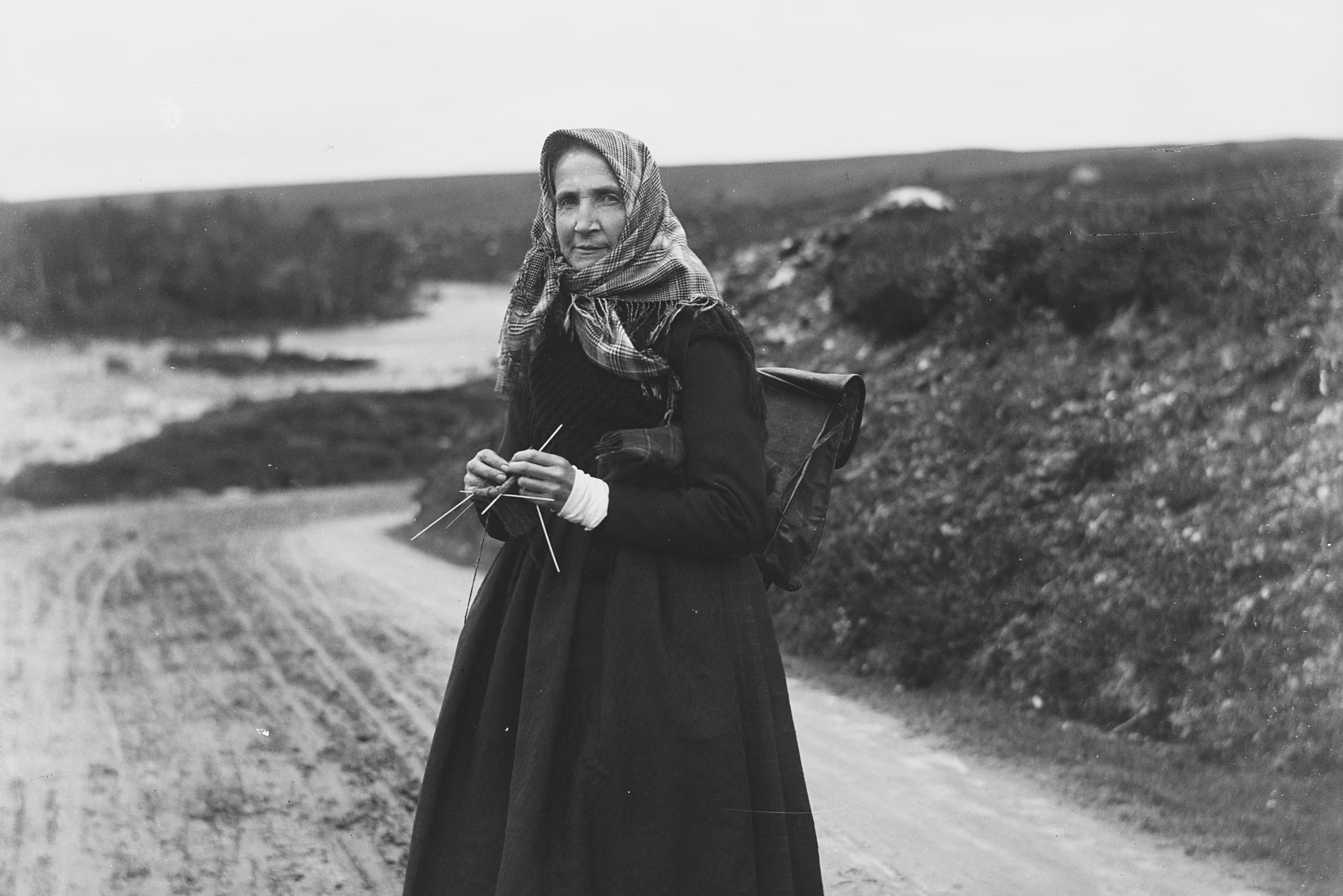 Svart- kvitt foto. Svartkledd kvinne med skreppe står midt i bilde med strikketøy.