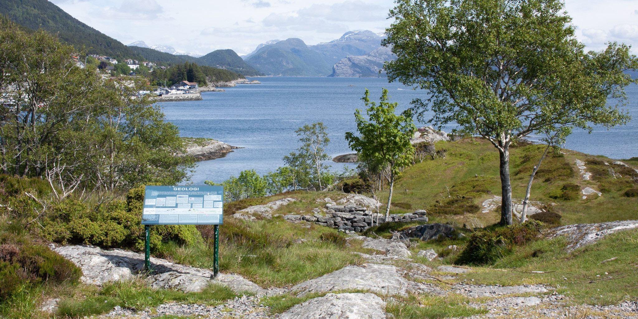 Utsikt over Brendøya, med hustufter, skilt om geografi og bjørketrær. Hav og fjell i bakgrunn.