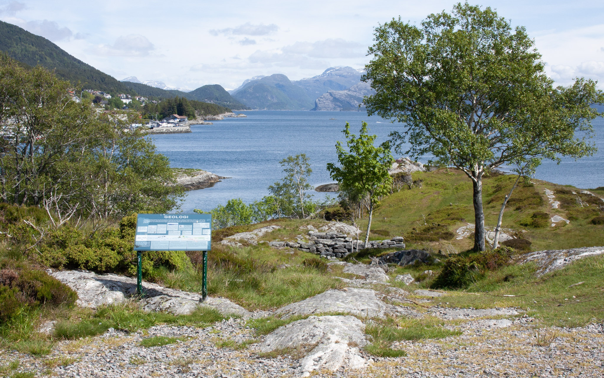 Utsikt over Brendøya, med hustufter, skilt om geografi og bjørketrær. Hav og fjell i bakgrunn.