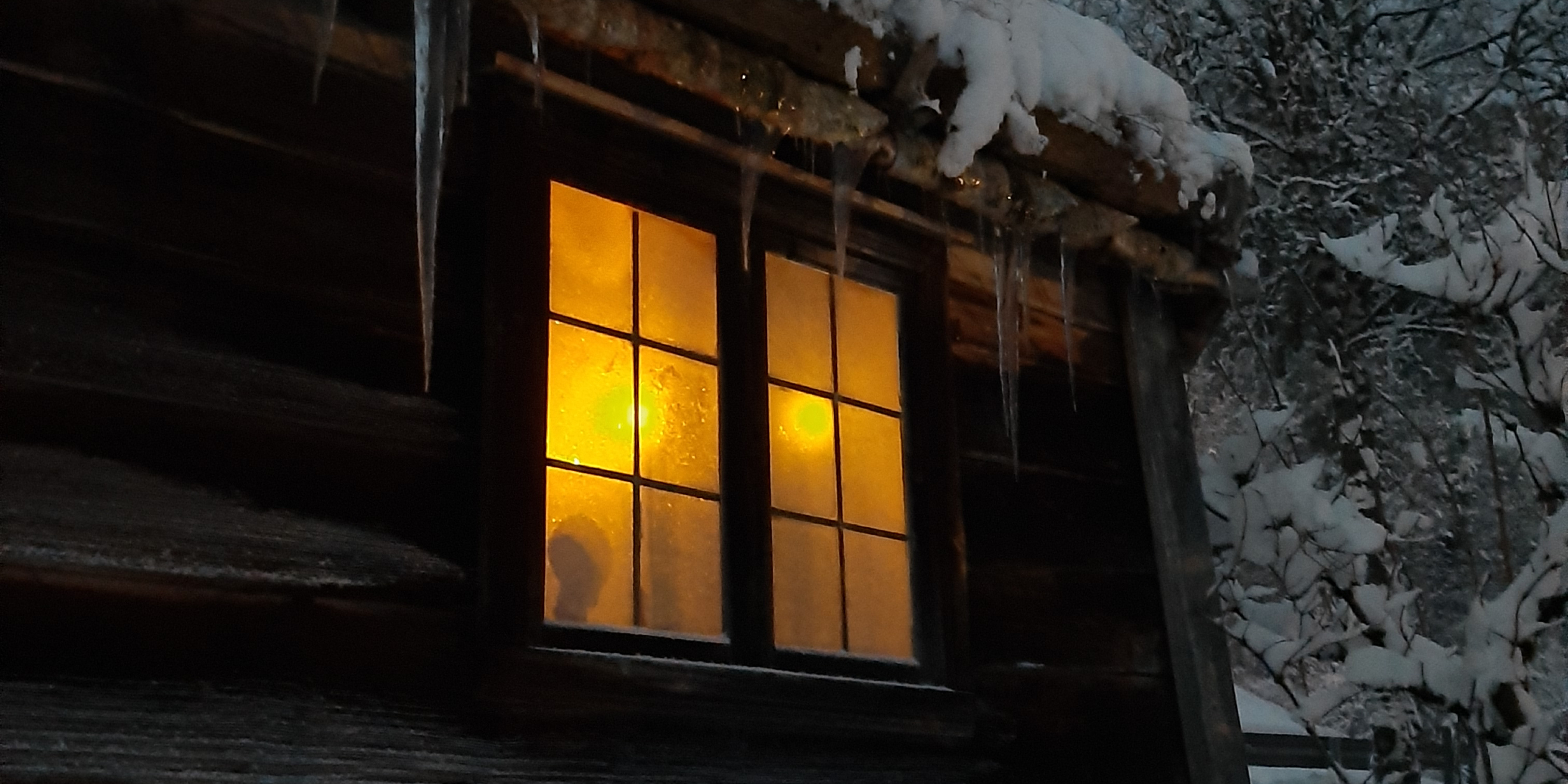 Utsnitt av lafta trehus med rimfrost på veggen. Snødekt tak med istappar hengande frå taket. Stearinlys i vindauget.